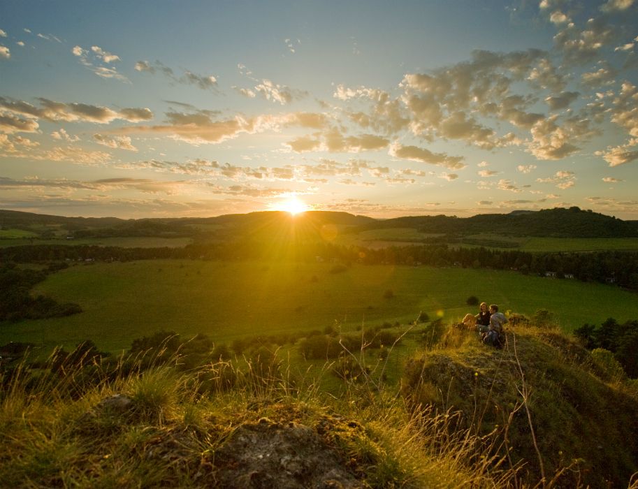 Sonnenuntergang über grüner Landschaft in der Eifel, mit Hügeln und Wolken. Zwei Personen sitzen auf einem Hügel im Vordergrund.