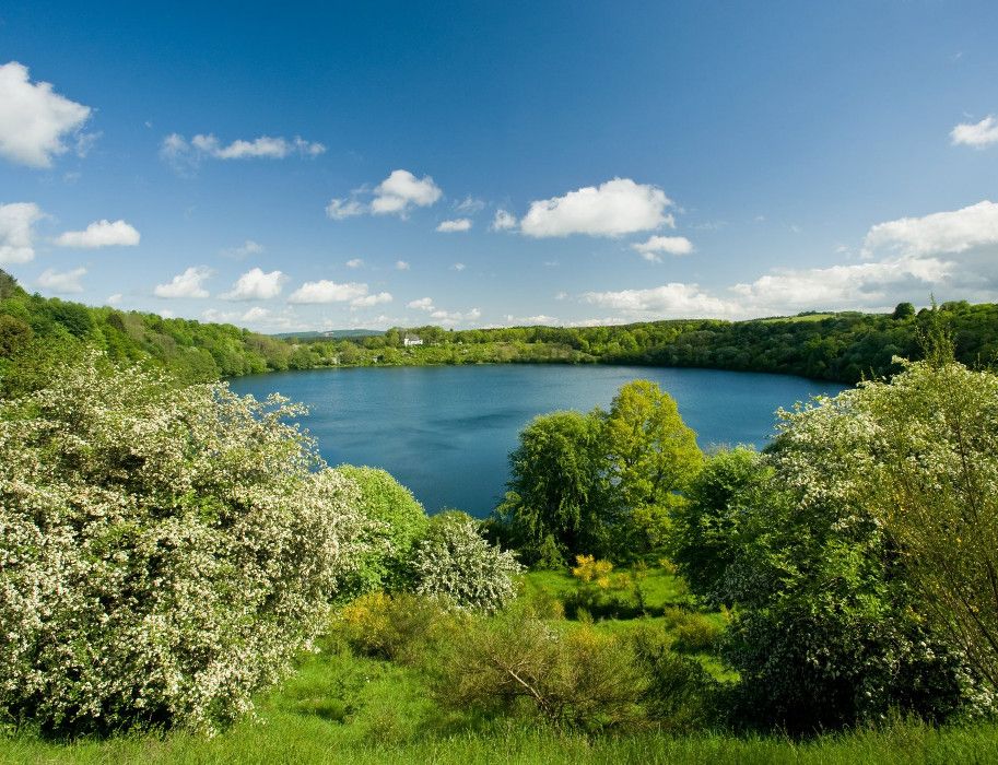 Weinfelder Maar in der Eifel mit blühenden Bäumen und blauem Himmel.