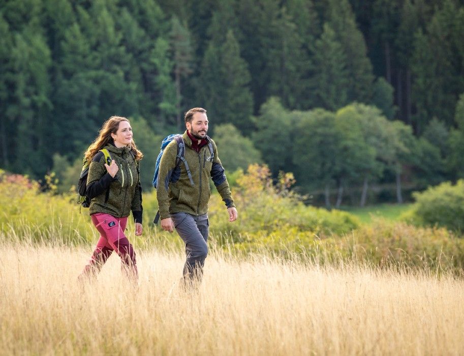 Zwei Wanderer mit Rucksäcken gehen durch eine Wiese, im Hintergrund dichter Wald. Sie tragen Outdoor-Kleidung und scheinen die Natur zu genießen.