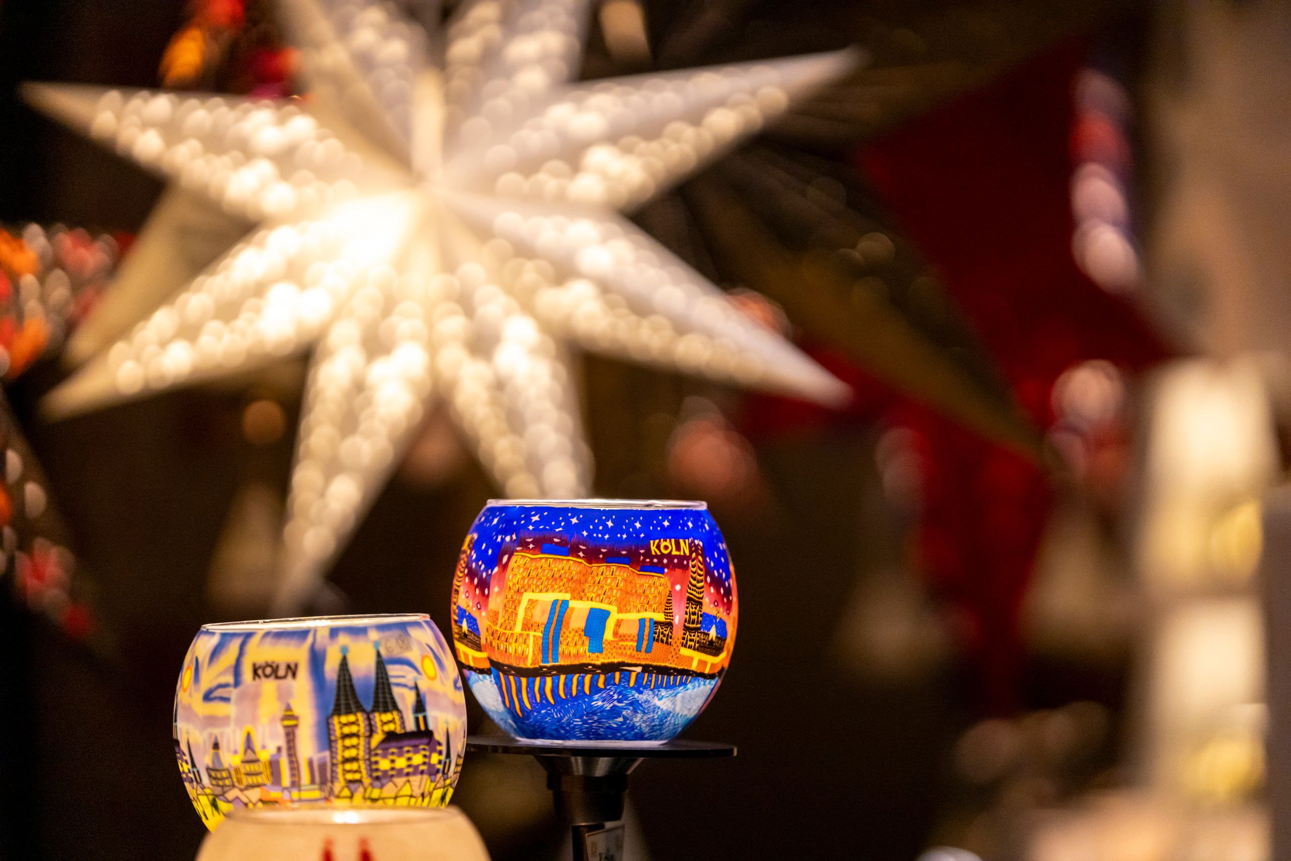 Bunte Kerzenhalter mit Kölner Motiven und im Hintergrund ein großer, leuchtender Stern sorgen für festliche Atmosphäre auf dem Markt der Engel.