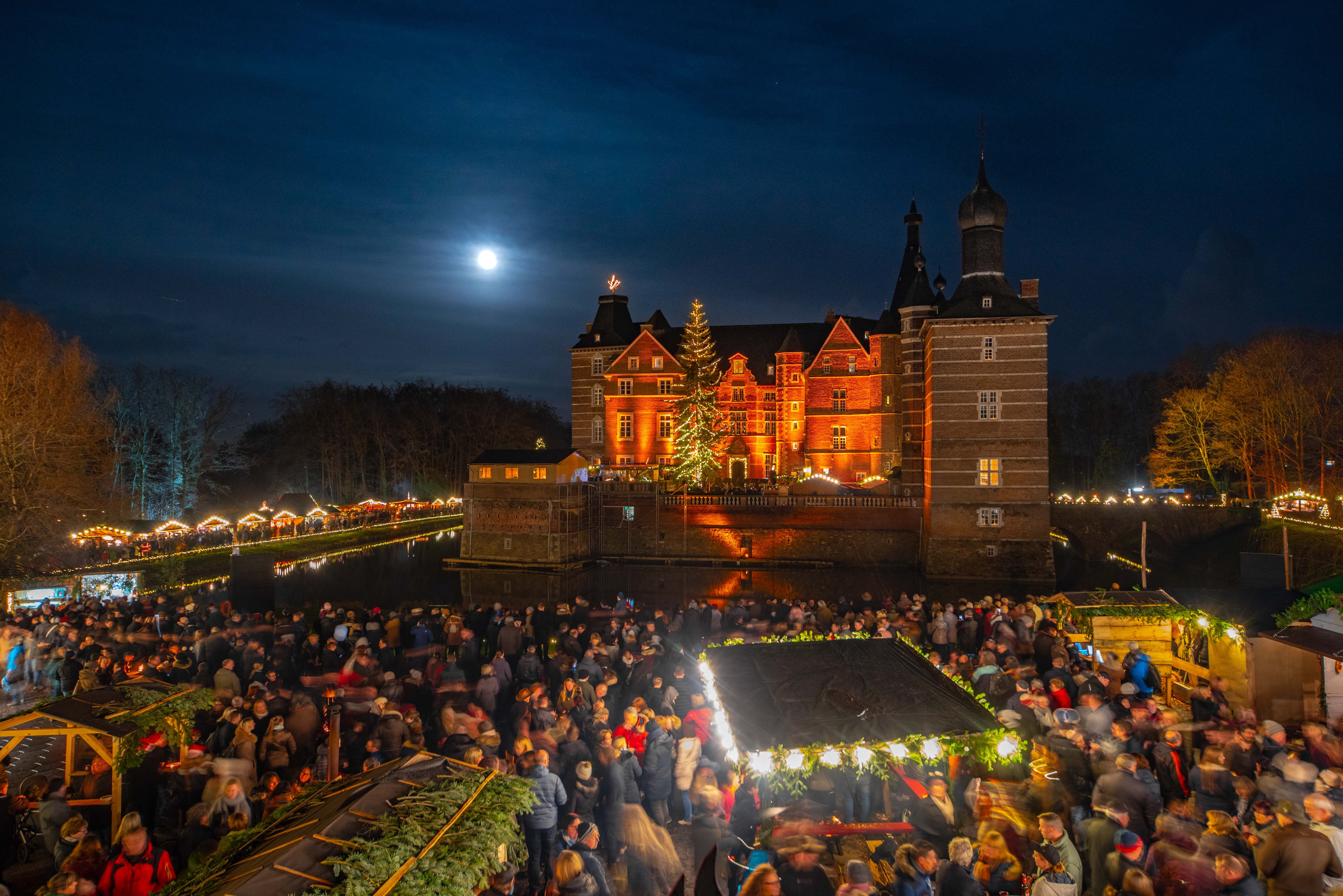 Schloss Merode bei Nacht, festlich beleuchtet mit Weihnachtsmarkt und zahlreichen Besuchern. Der Vollmond scheint am Himmel.