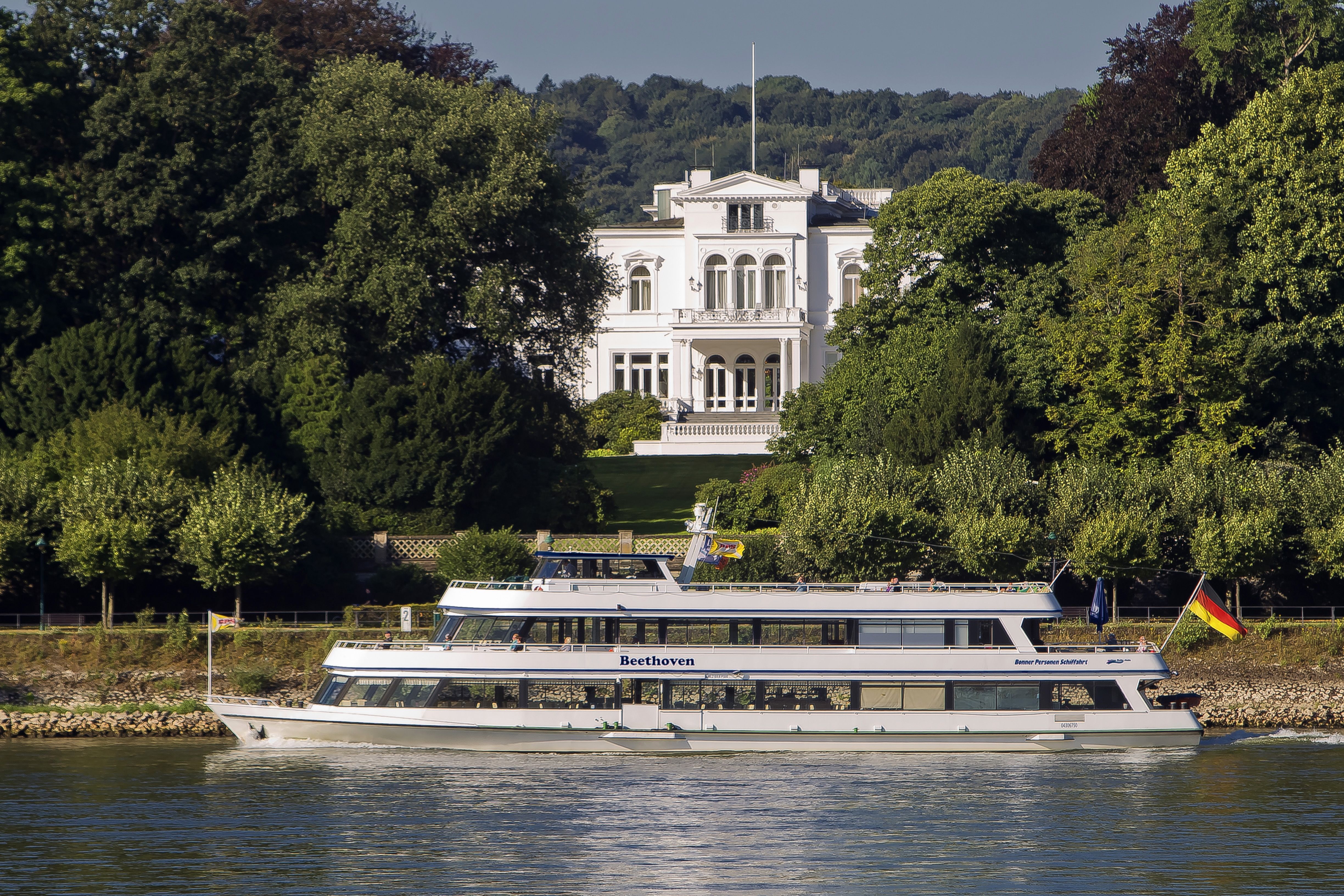 Die Villa Hammerschmidt am Rhein, umgeben von Bäumen. Ein Ausflugsschiff namens Beethoven fährt auf dem Fluss entlang.
