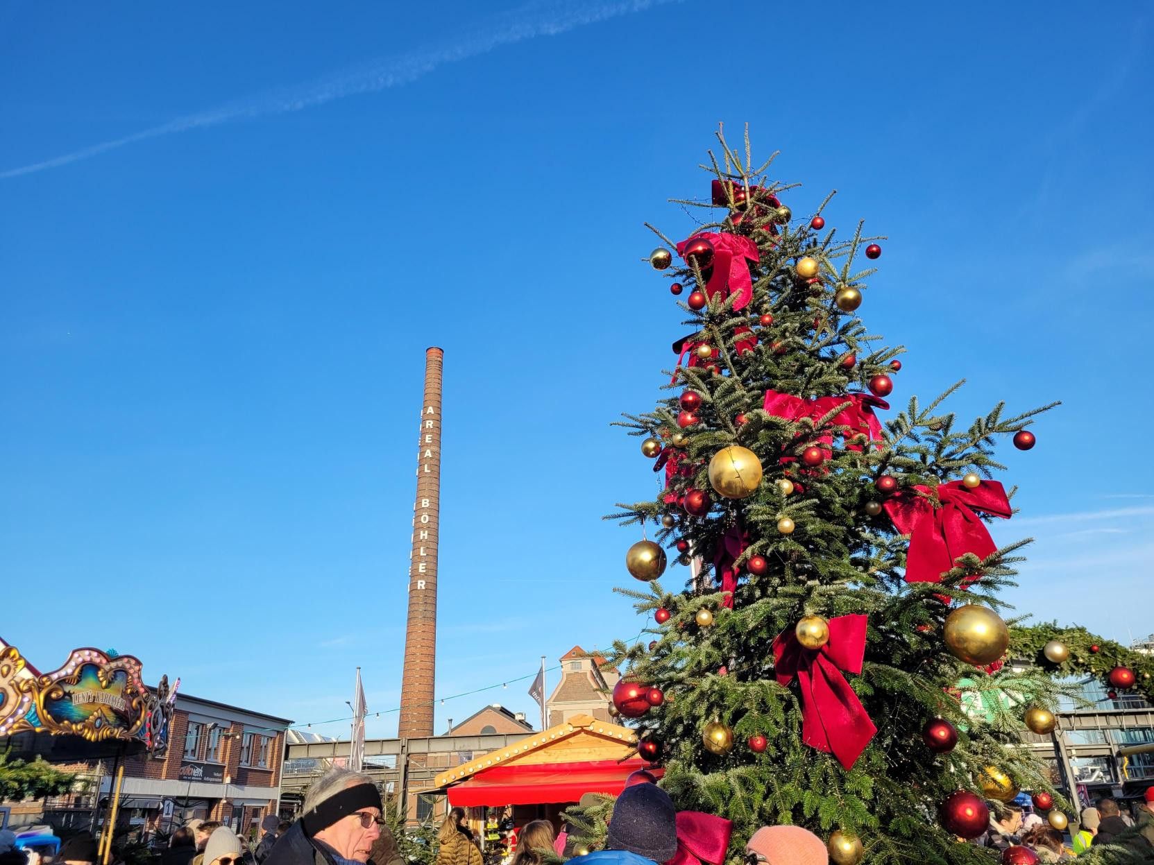 Großer Weihnachtsbaum mit roten Schleifen und goldenen Kugeln, umgeben von Menschen. Im Hintergrund der Schornstein 'Areal Böhler' unter blauem Himmel.