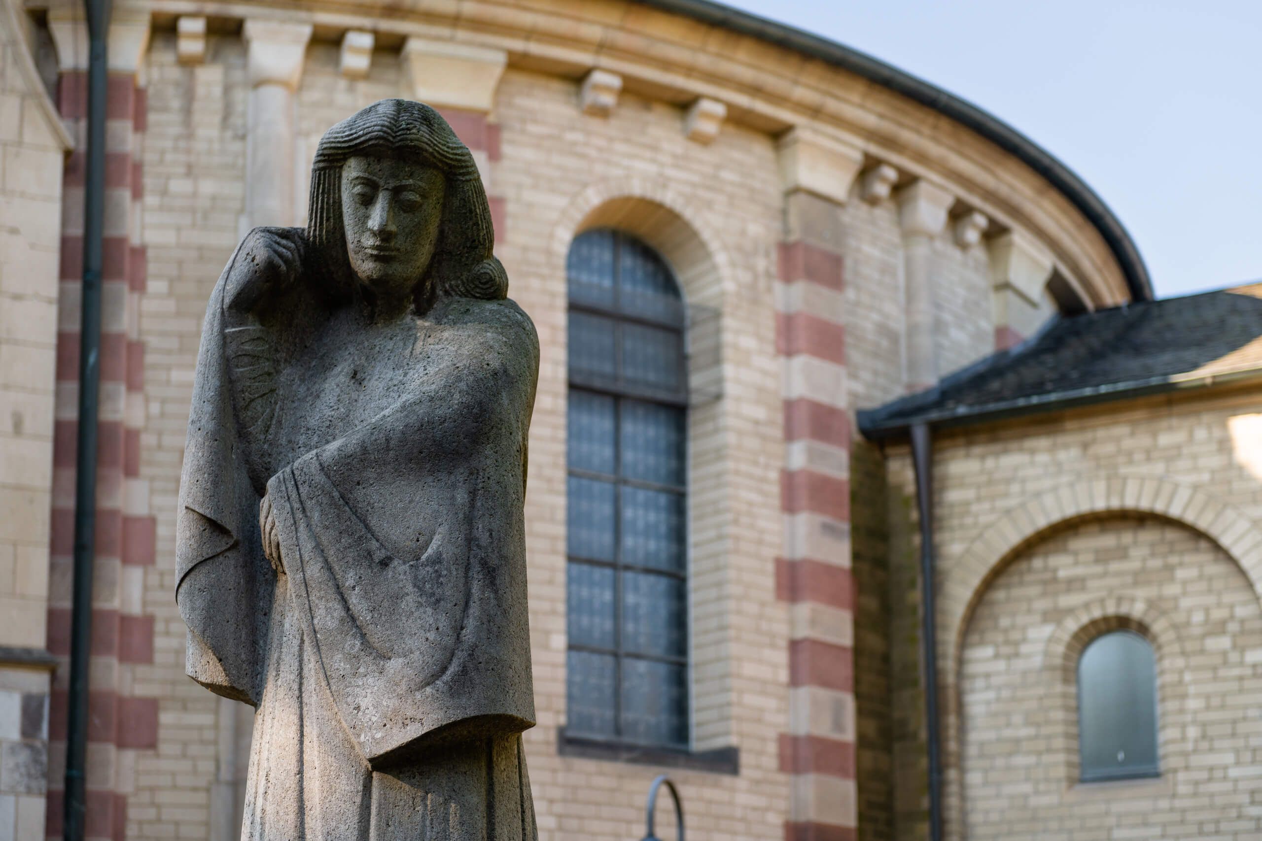 Eine steinerne Statue steht vor der romanischen Kirche Sankt Maria im Kapitol in Köln, mit charakteristischen Rundbogenfenstern im Hintergrund.