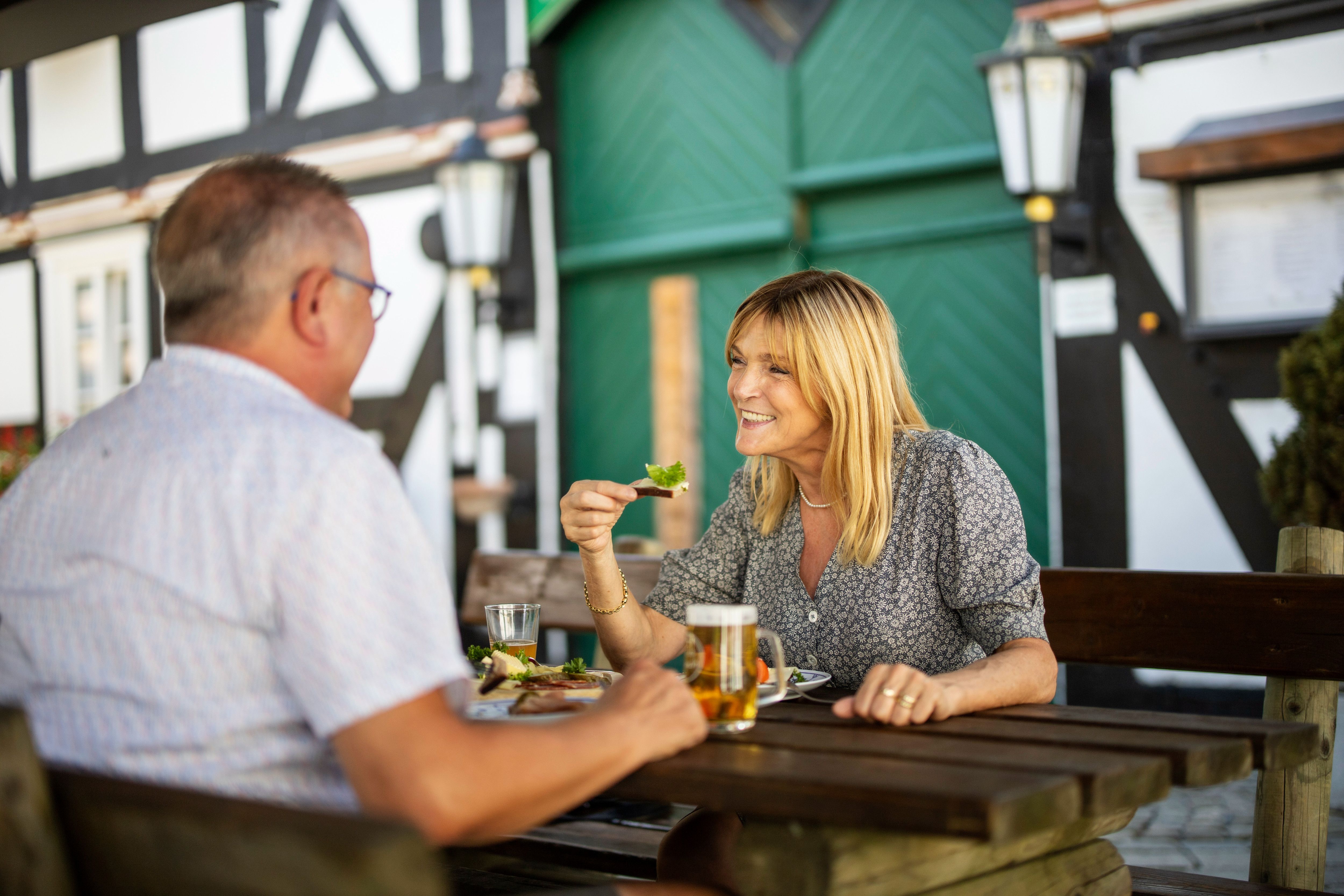 Ein Mann und eine Frau essen gemeinsam an einem Holztisch im Freien vor einem Fachwerkhaus.
