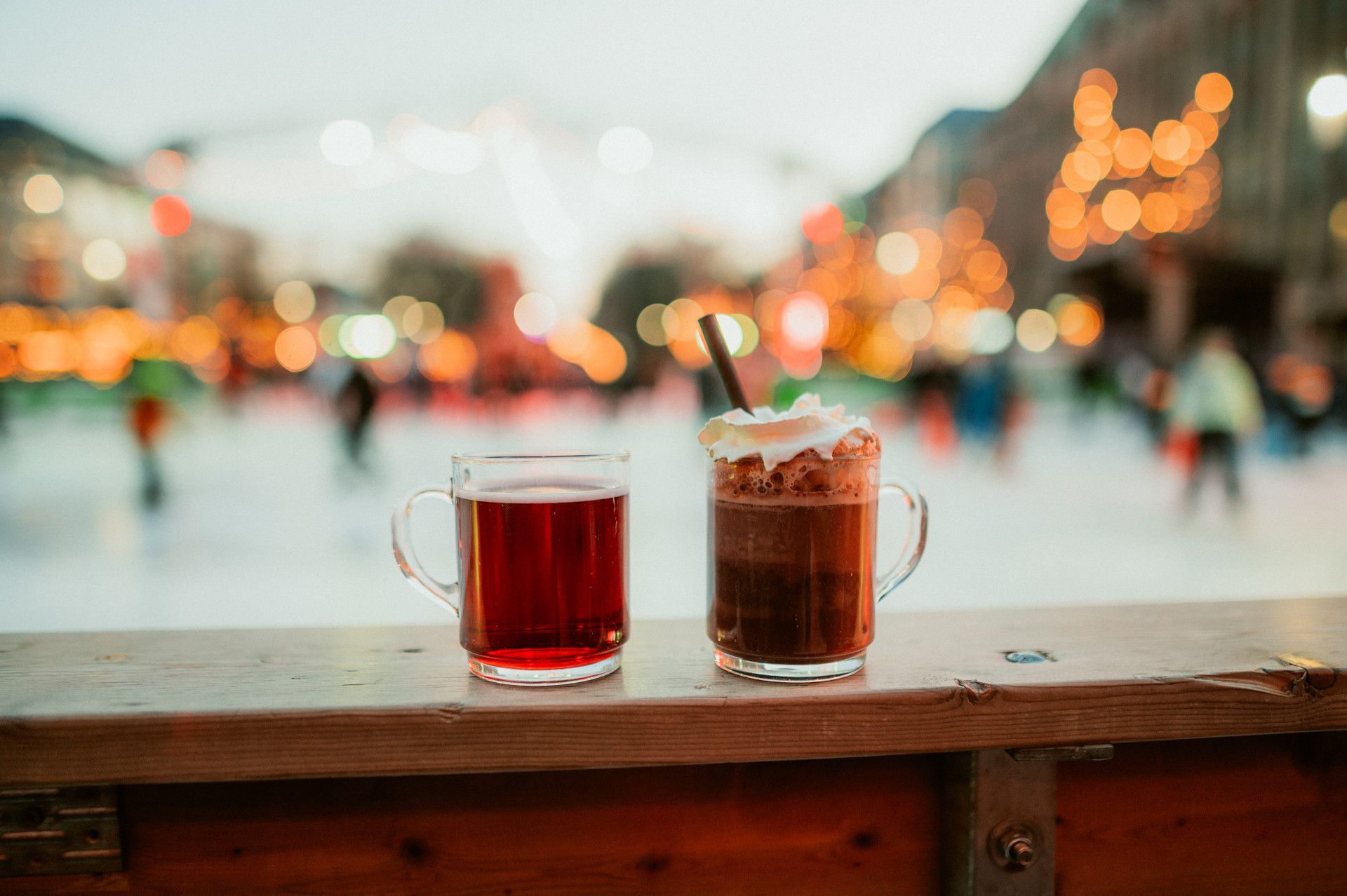 Zwei Gläser mit Glühwein und heißer Schokolade auf einem Holztisch, im Hintergrund eine verschwommene Eislaufbahn mit Lichtern.