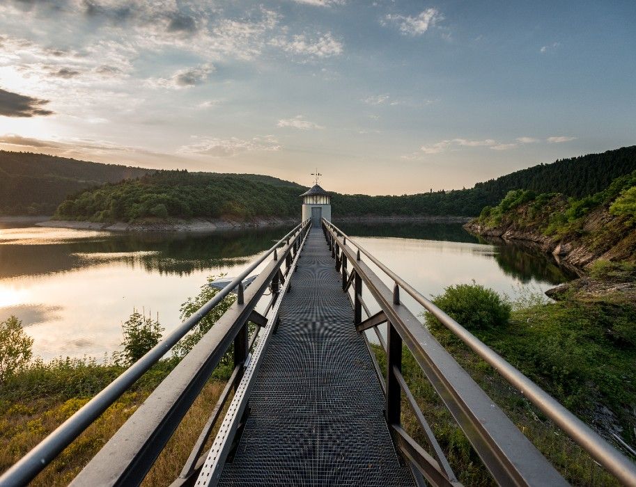 Eine Metallbrücke führt zur Urfttalsperre, umgeben von ruhigem Wasser und grünen Hügeln bei Sonnenuntergang.