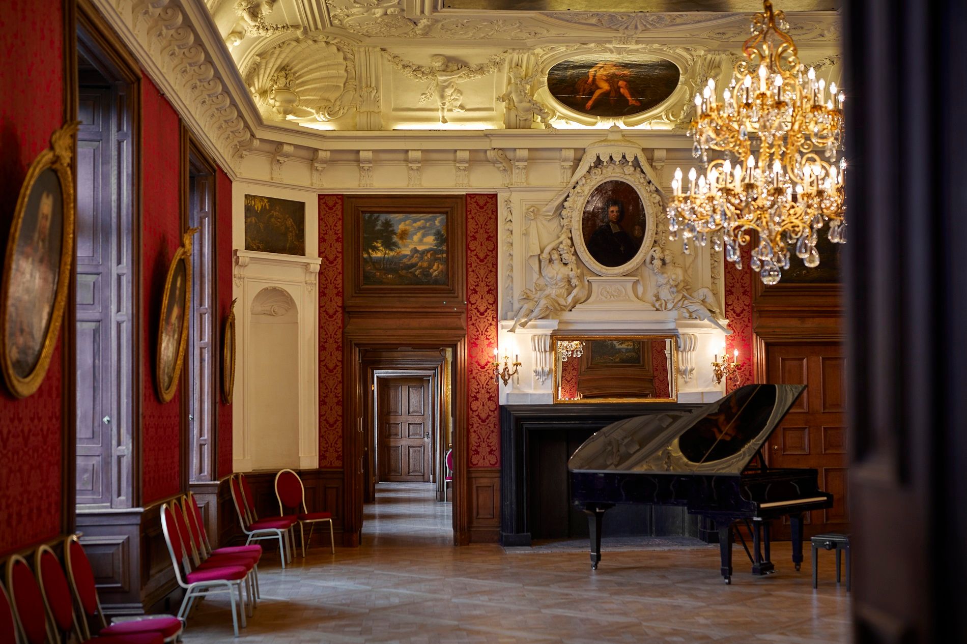 Prunkvoller Raum im Schloss Nordkirchen mit roter Tapete, Gemälden, einem Kronleuchter und einem schwarzen Flügel.