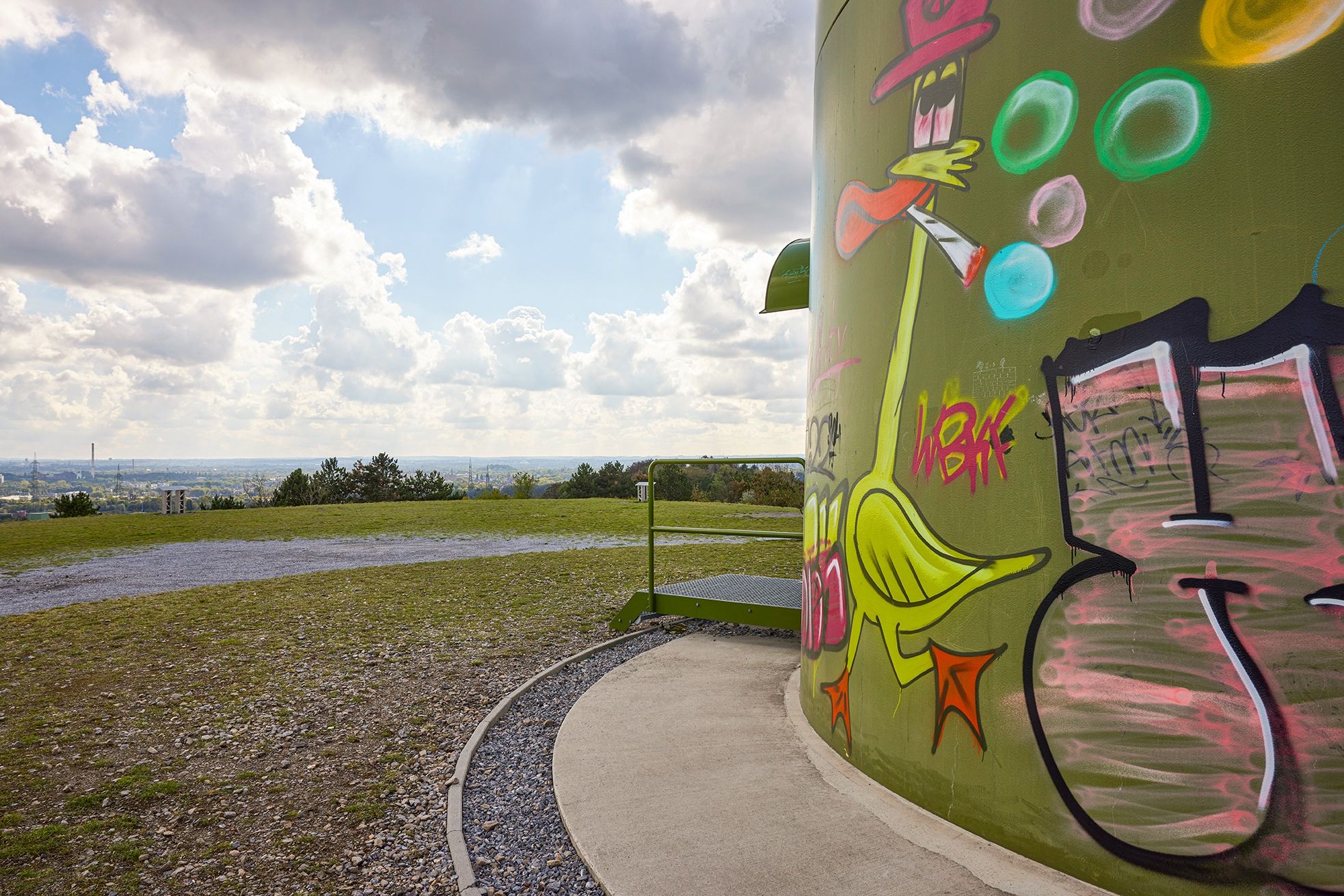 Graffiti auf einem grünen Turm mit Landschaft im Hintergrund.