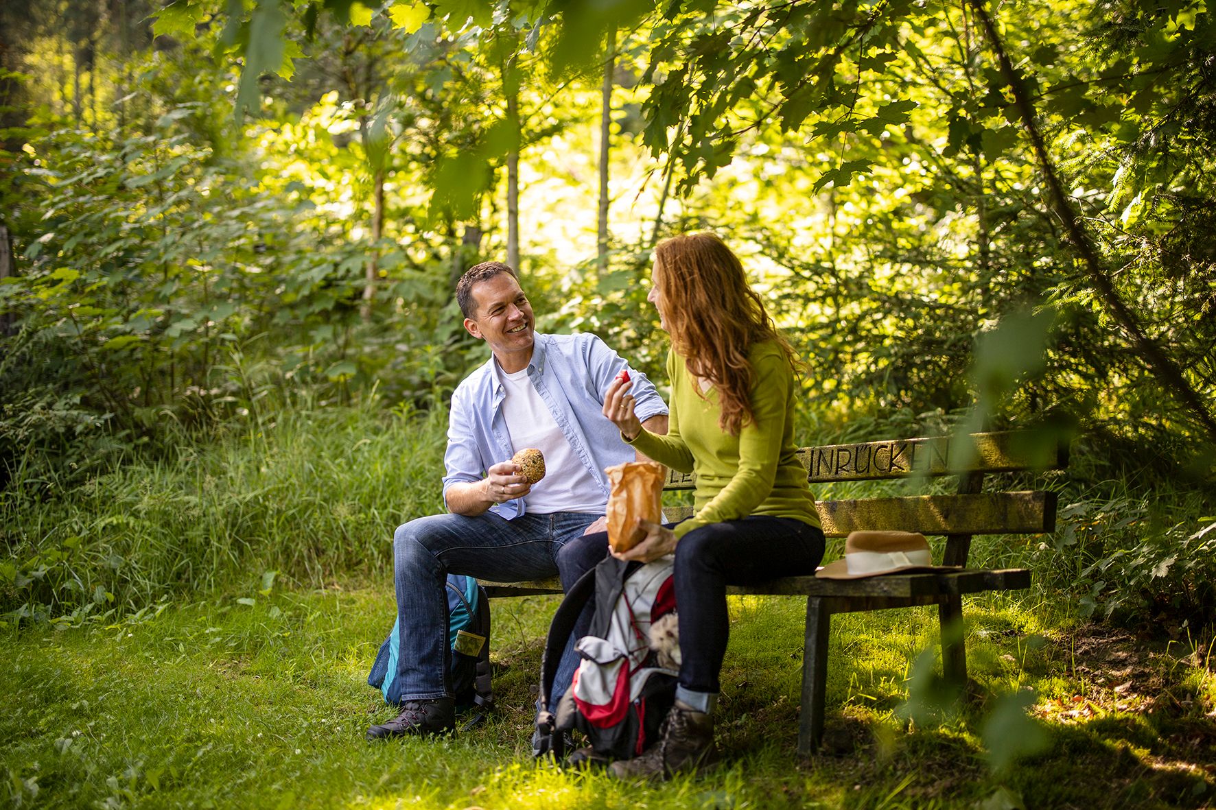 Zwei Personen sitzen auf einer Bank im Grünen, trinken etwas und unterhalten sich entspannt im Wald.