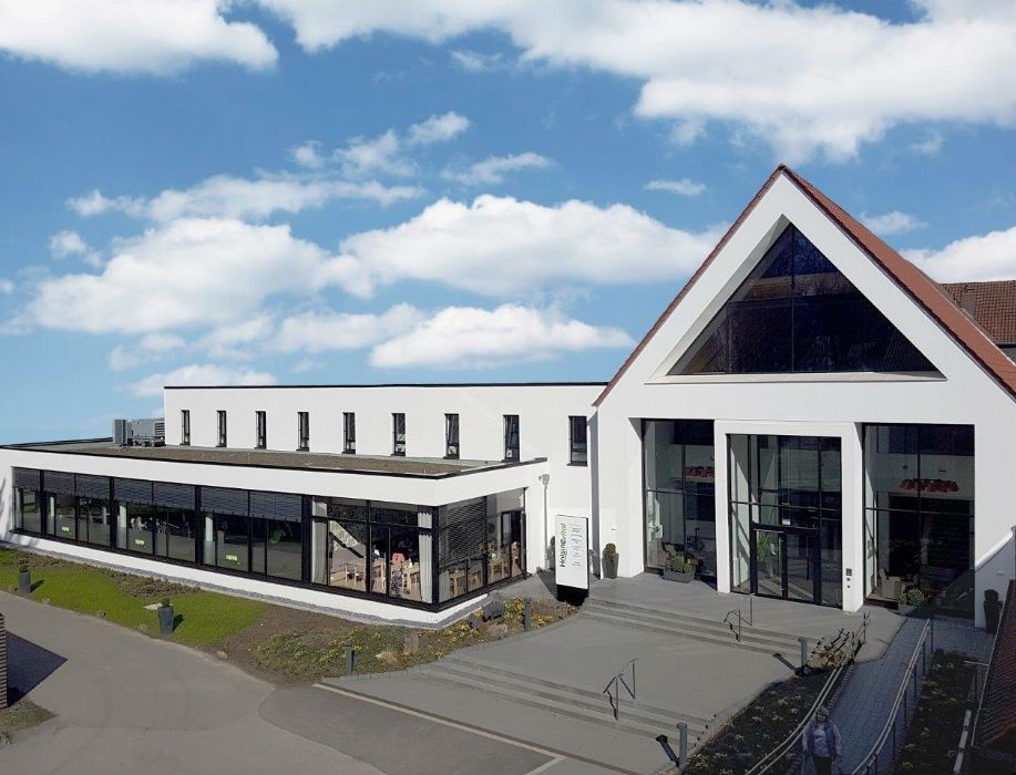 Moderner Gebäudeeingang mit großen Glasfenstern und dreieckigem Giebel unter blauem Himmel.