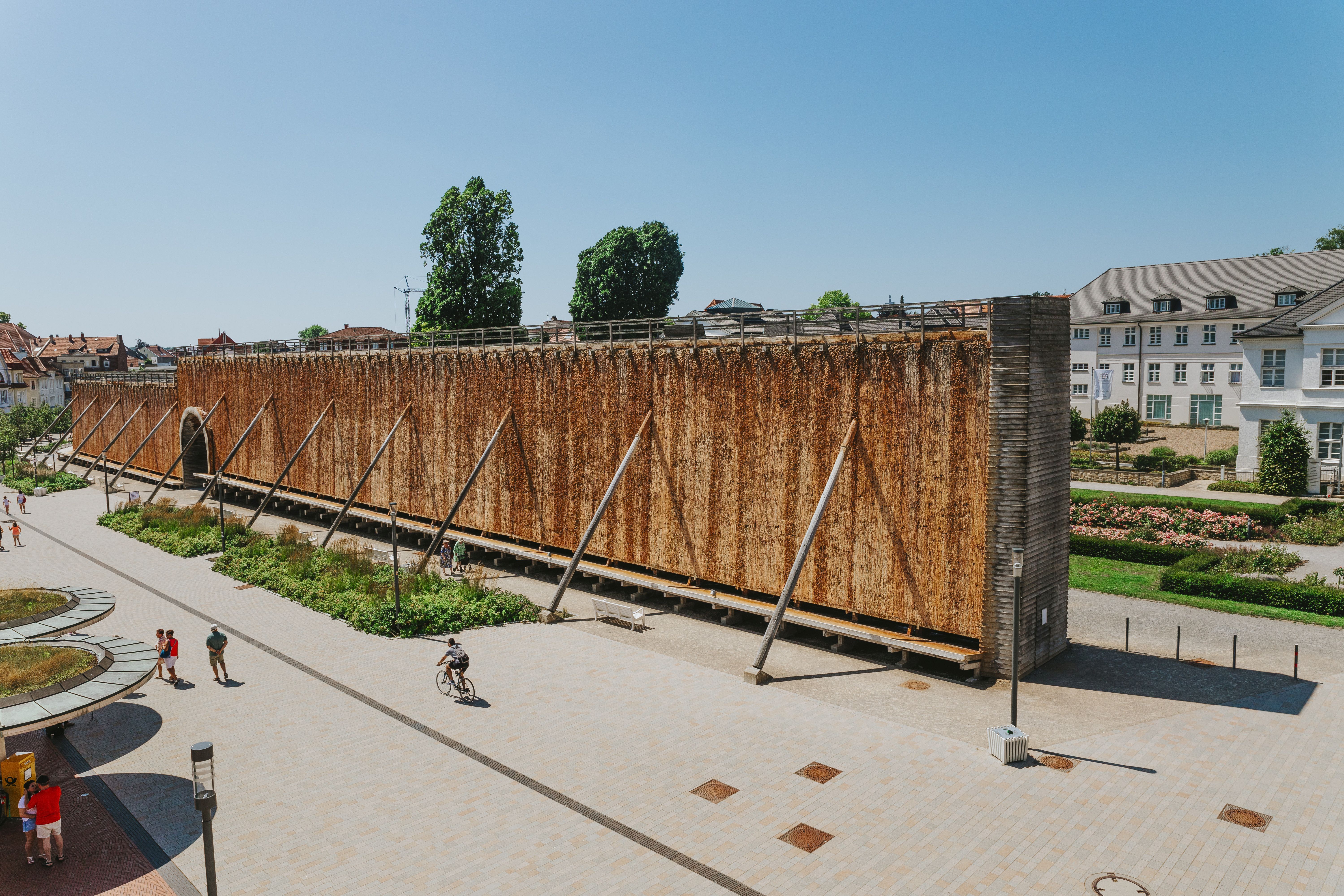 Ein Gradierwerk im Rosengarten mit Spaziergängern und einem Radfahrer auf einem sonnigen Platz.
