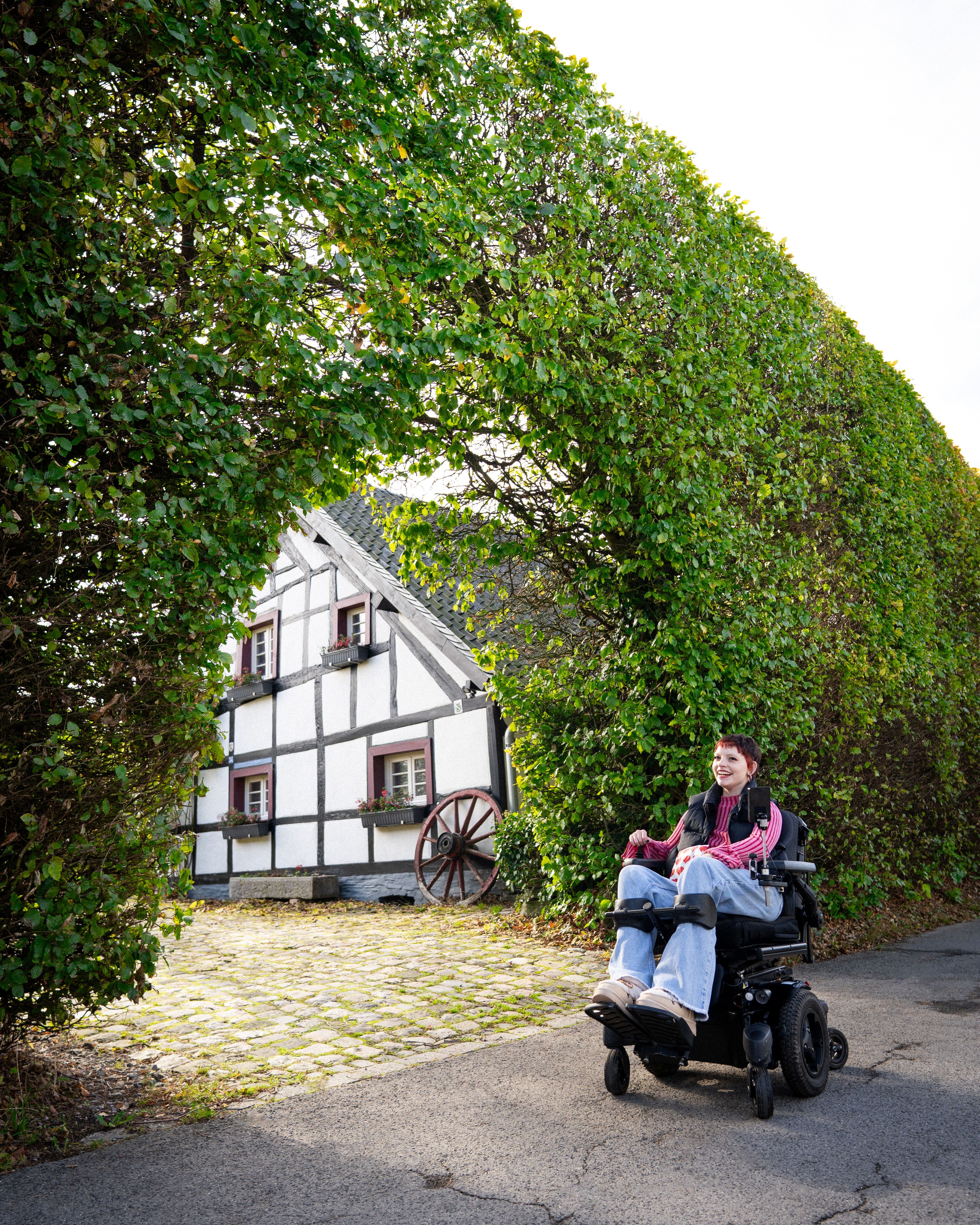 Eine Rollstuhlfahrerin strahlt vor Freude vor einer Haushecke mit Blick auf ein Fachwerkhaus in Monschau-Höfen