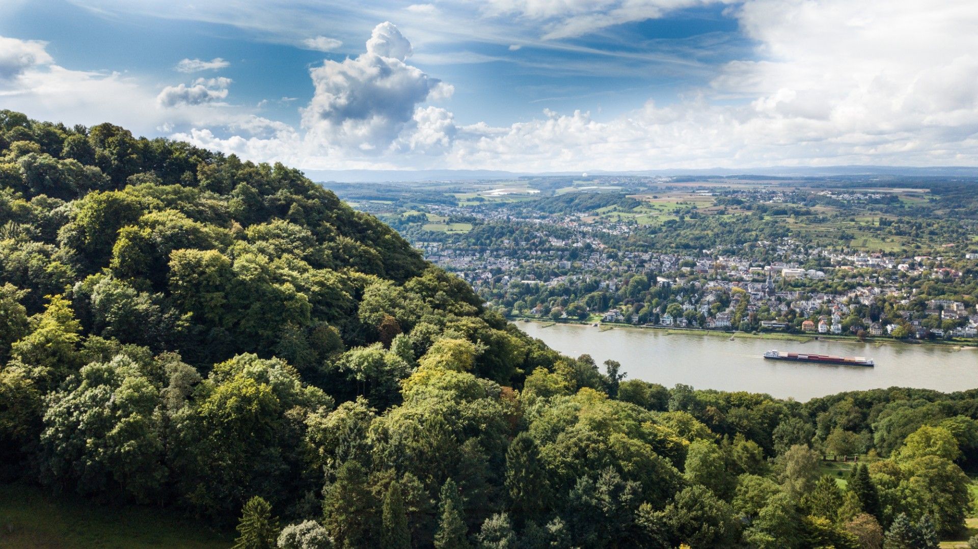 Luftaufnahme des Rheintals bei Königswinter mit bewaldetem Hügel im Vordergrund und Stadt im Hintergrund.