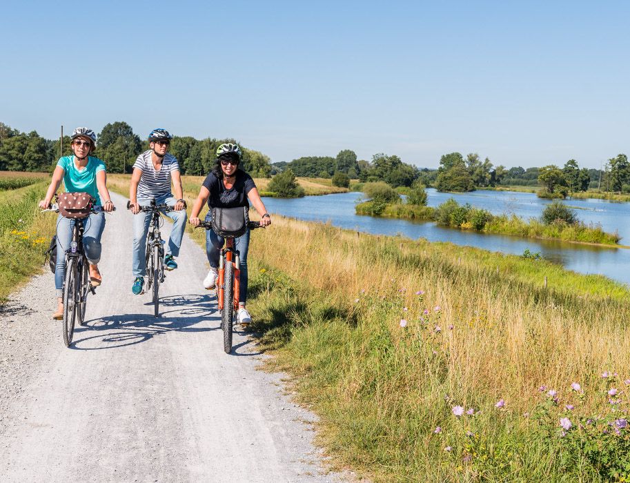 Drei Personen fahren auf einem Schotterweg mit Fahrrädern entlang eines Flusses im Steinhorster Becken.