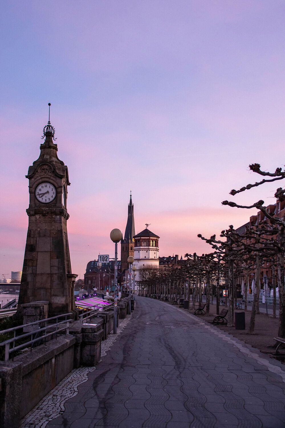 Rheinuferpromenade in Düsseldorf bei Sonnenuntergang. Ein Uhrturm steht links, Bäume säumen den Weg. Der Himmel ist rosa gefärbt.