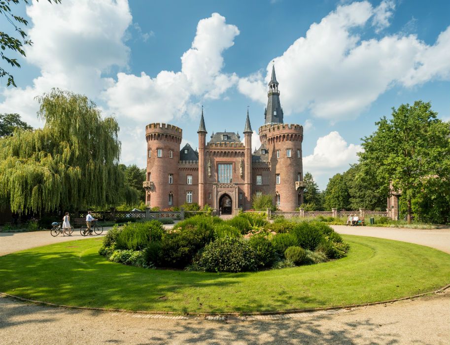 Schloss Moyland mit zwei markanten Türmen, umgeben von einem gepflegten Garten und Spaziergängern auf einem sonnigen Tag