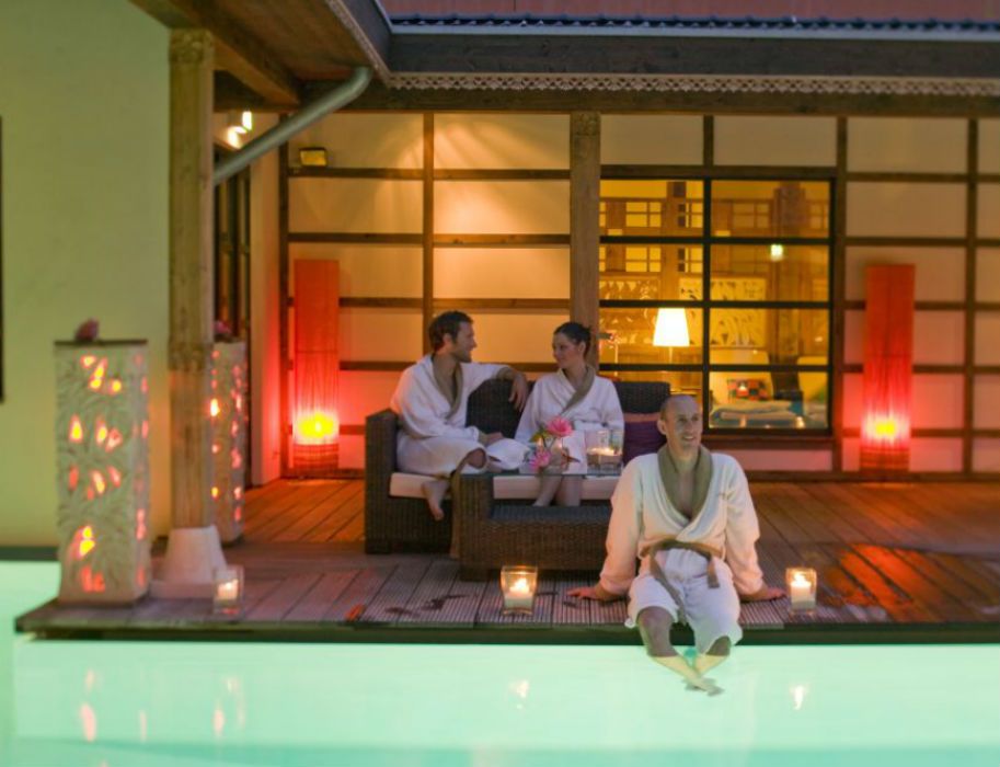 Drei Personen in Bademänteln entspannen im Bali Therme Atrium neben einem Pool.