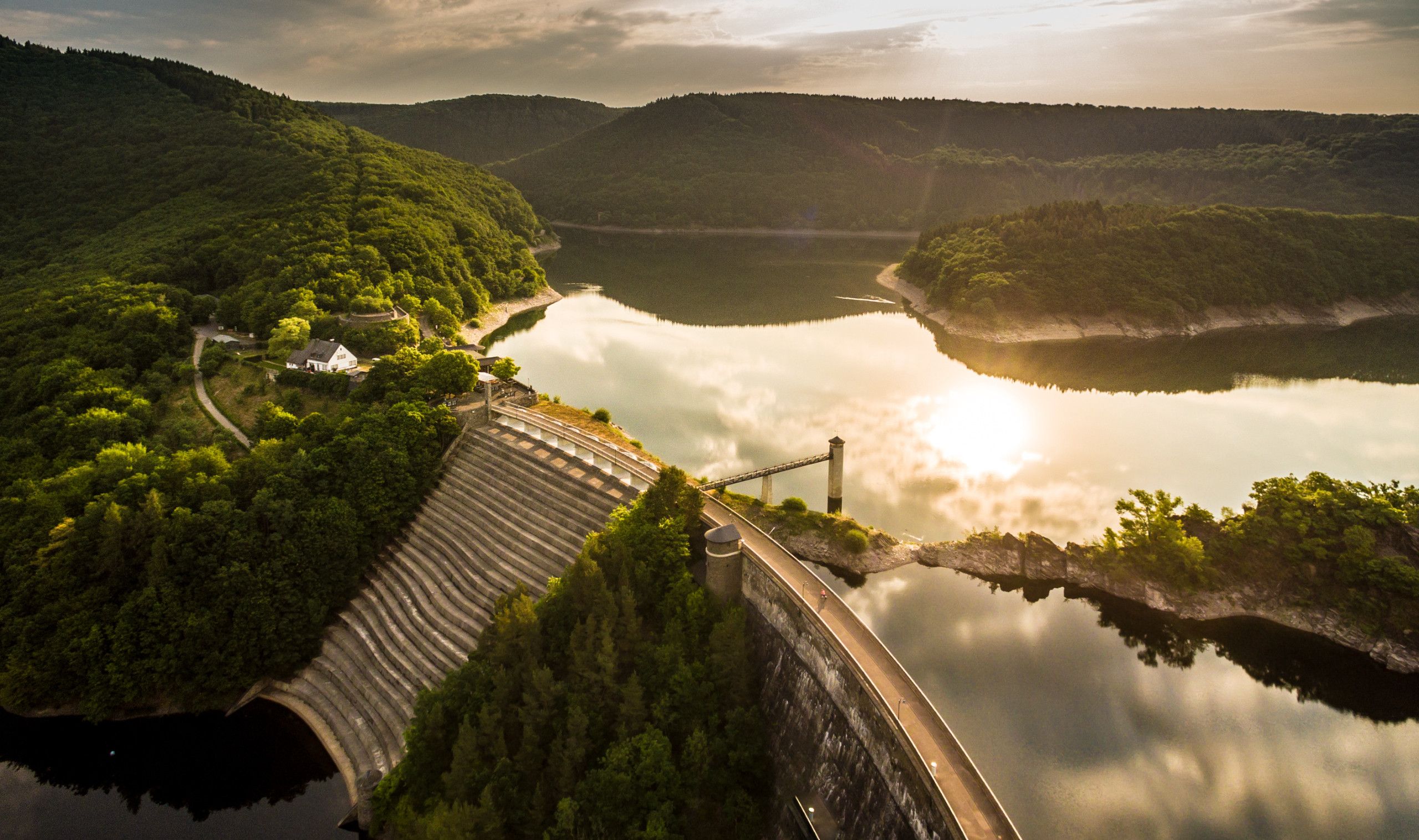 Luftaufnahme der Urfttalsperre mit Stausee und bewaldeten Hügeln im Sonnenuntergang.