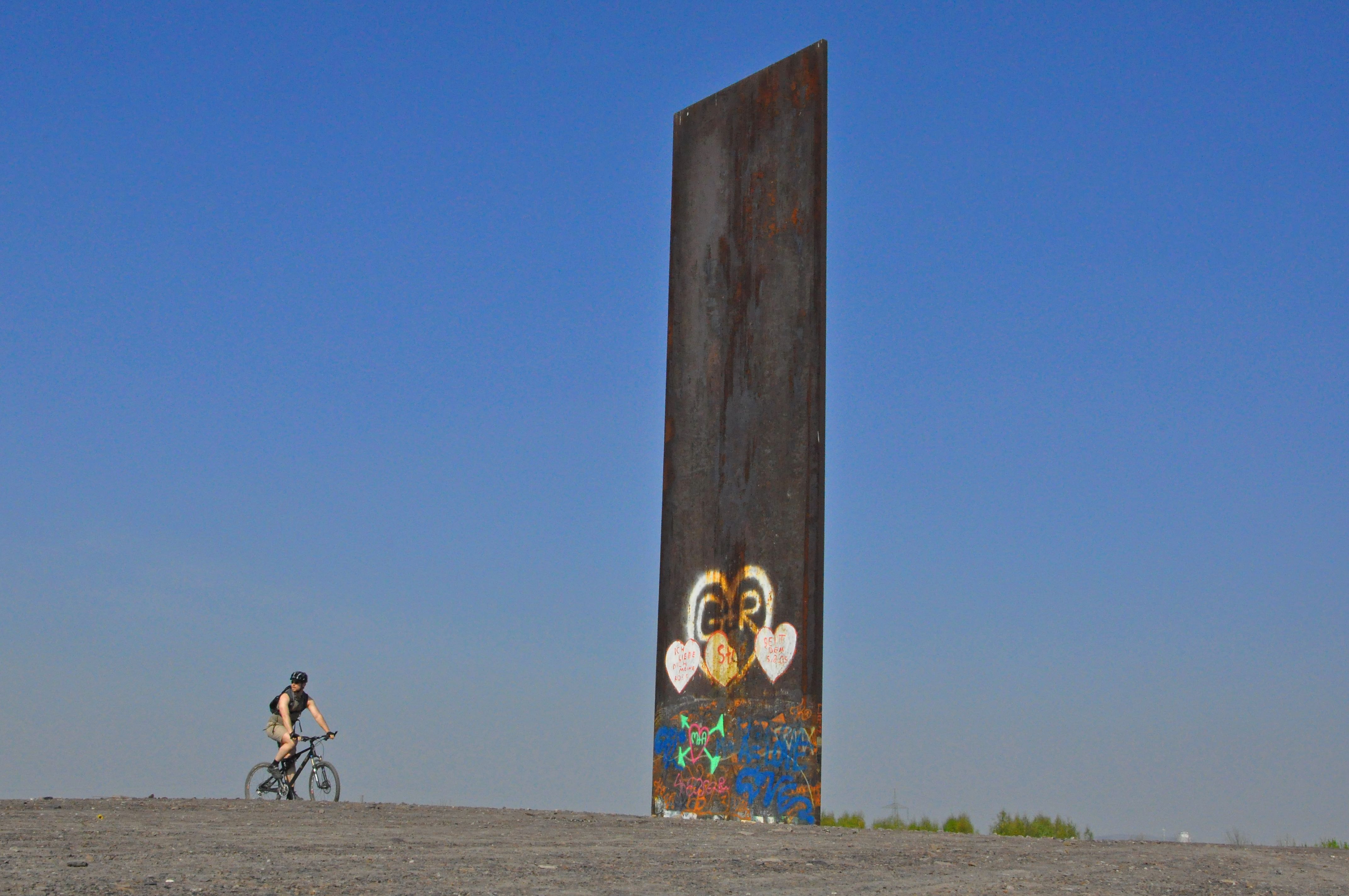Ein Radfahrer fährt an einer großen, bemalten Stahlplatte unter klarem, blauem Himmel.