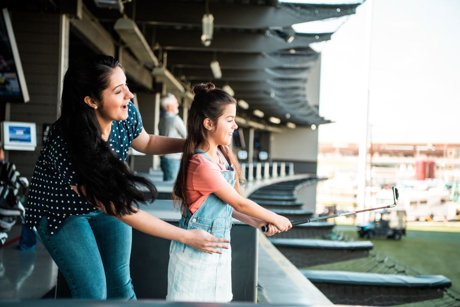 Eine Frau hilft einem Mädchen beim Golfspielen in einer Topgolf-Anlage.