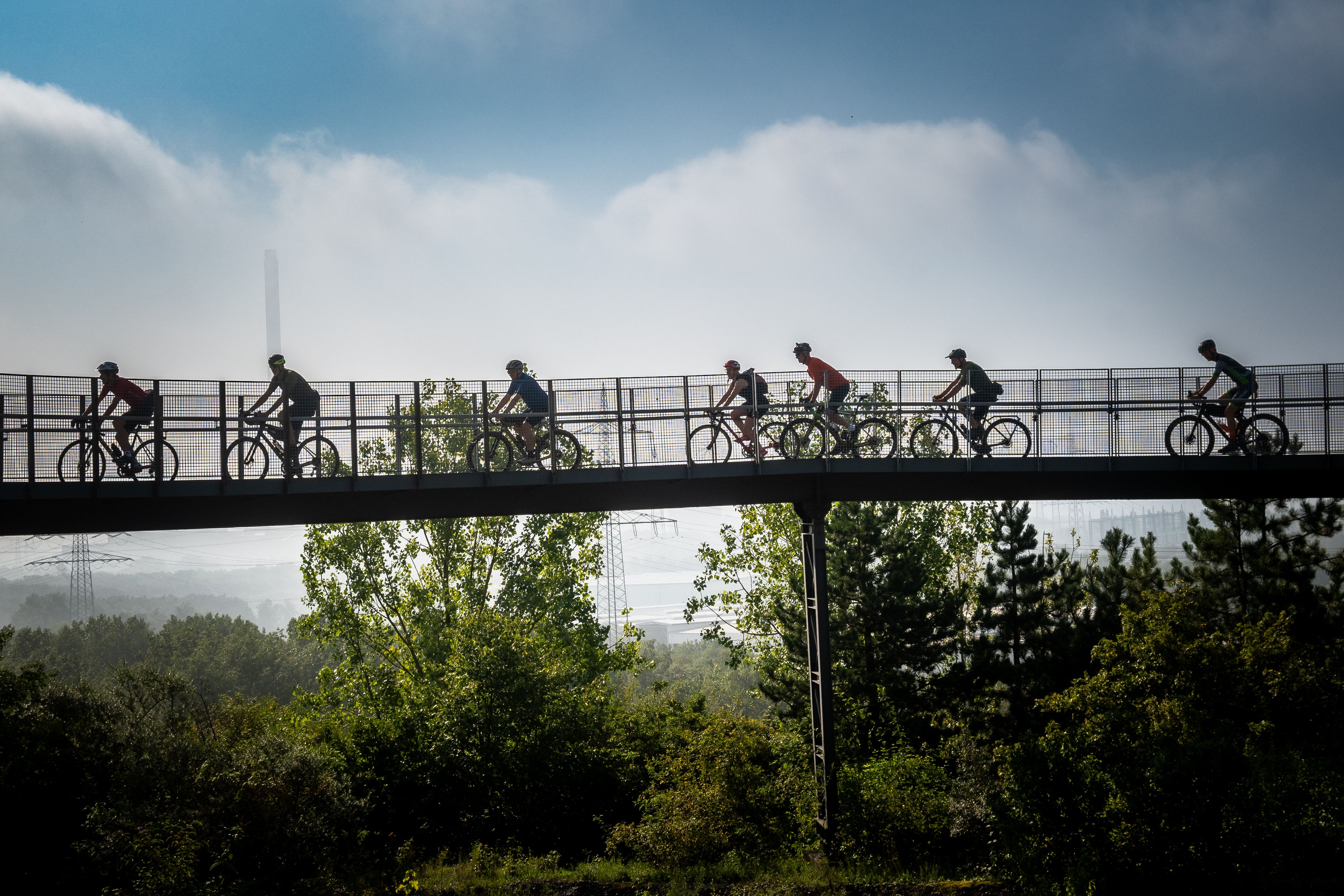 Silhouette von Radfahrern auf einer Brücke vor bewölktem Himmel.