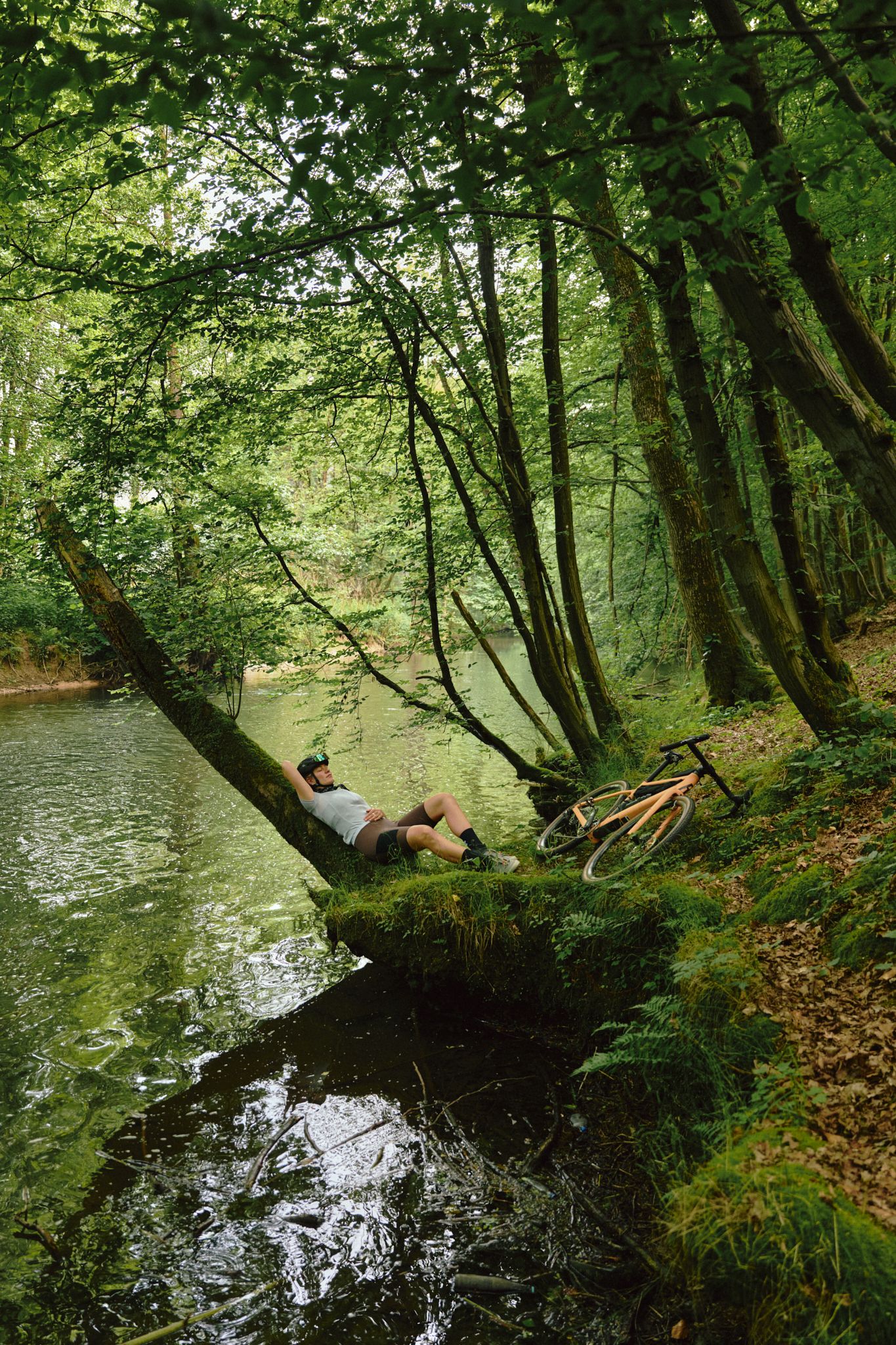 Person entspannt sich auf einem Baumstamm am Flussufer im Wald, neben einem Fahrrad.
