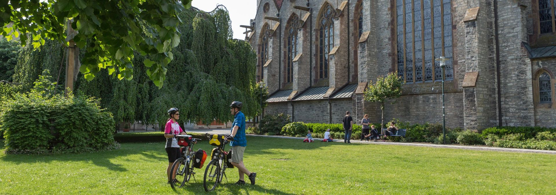 Zwei Radfahrer stehen vor einer Kirche im Grünen, umgeben von Bäumen und Menschen, die sich entspannen. Am dritten oder vierten Reisetag können Radreisende die Sehenswürdigkeiten im Osnabrücker Land ausmachen.