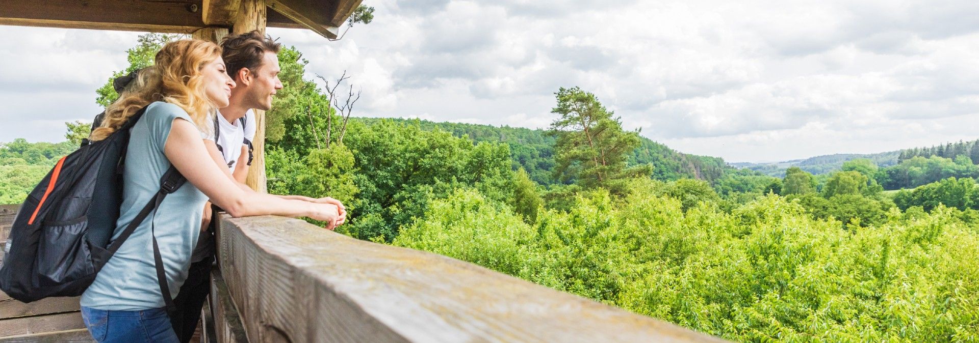Zwei Personen genießen die Aussicht von einem Holzturm auf einen grünen Wald unter bewölktem Himmel.