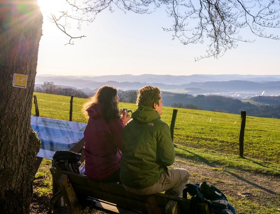 Zwei Wanderer sitzen auf einer Bank im Sauerland und genießen die Aussicht auf Hügel und Täler bei Sonnenuntergang.