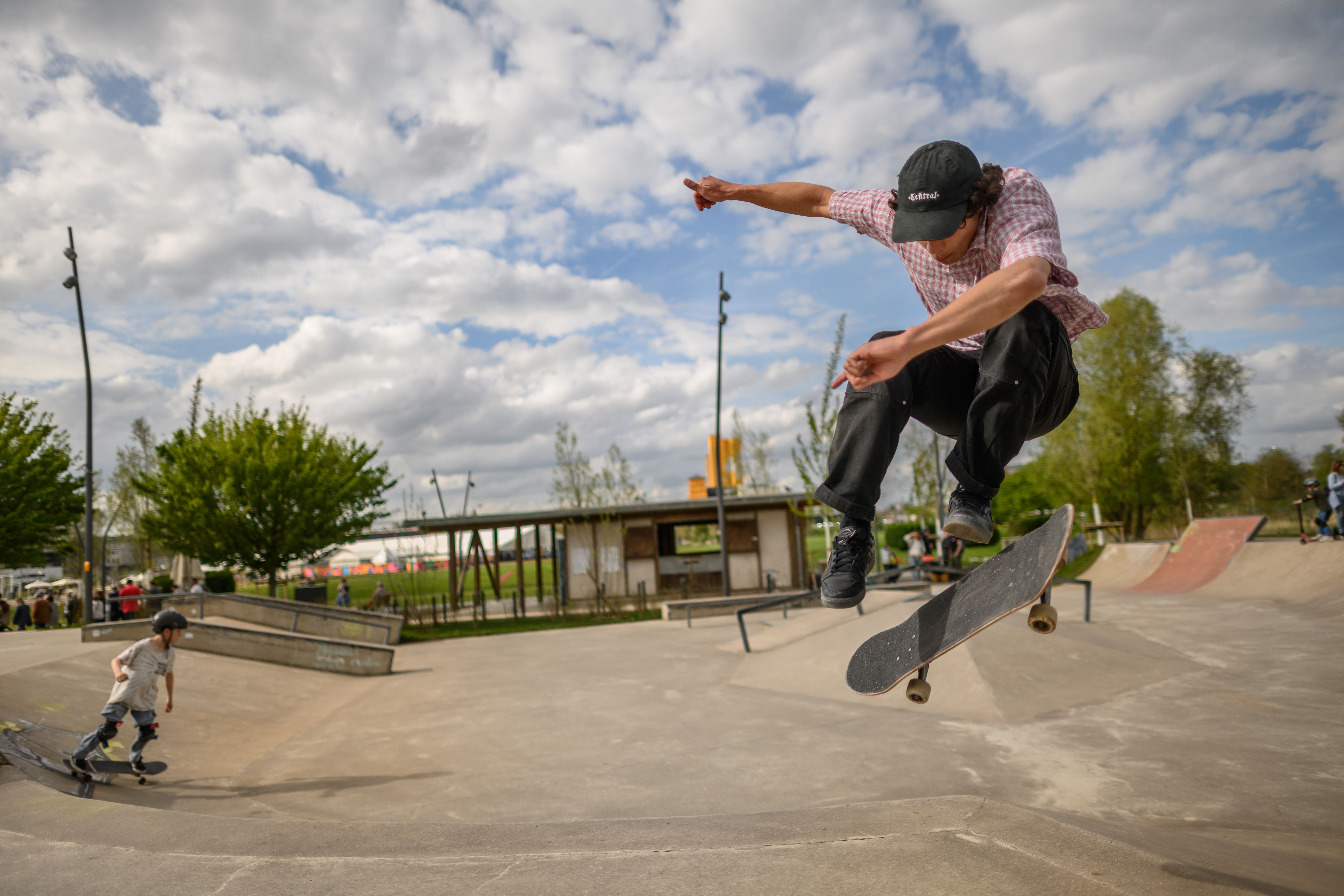 Ein Skateboarder führt einen Trick in einem Skatepark aus, während ein Kind auf einem Skateboard im Hintergrund zu sehen ist.