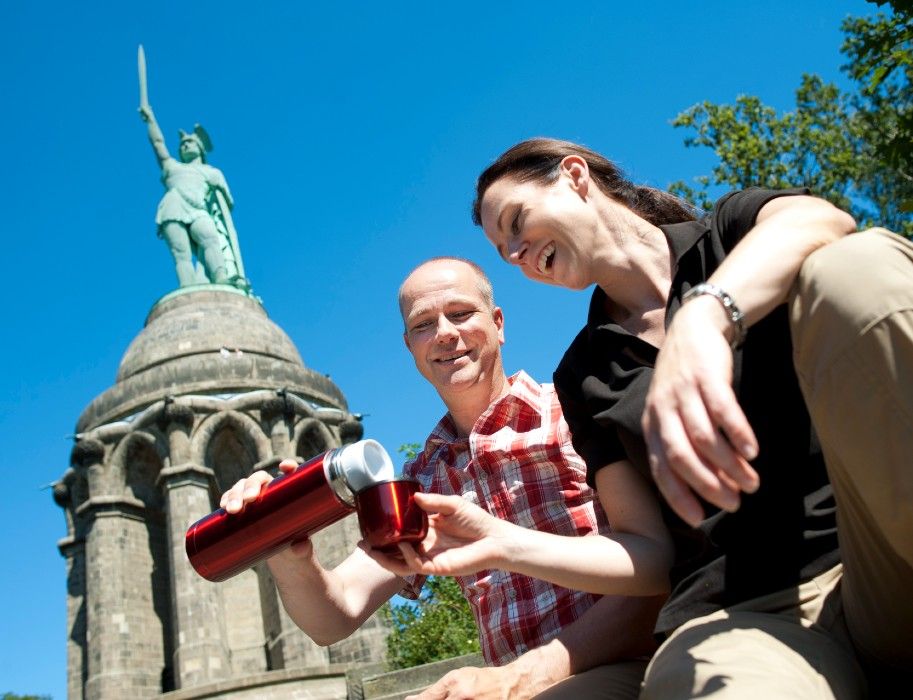 Zwei Personen sitzen vor dem Hermannsdenkmal und gießen aus einer roten Thermoskanne ein Getränk ein. Der Himmel ist klar und blau.
