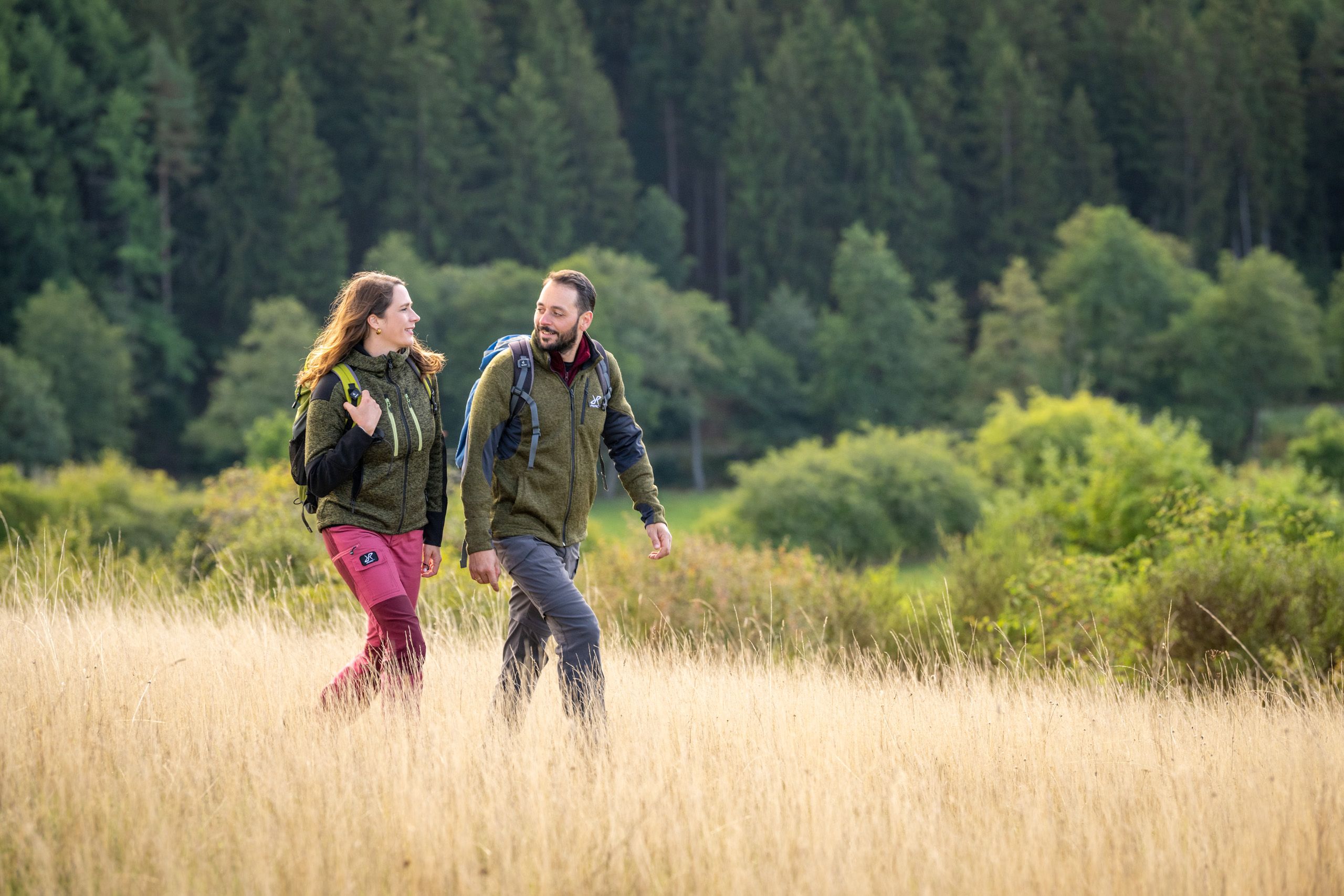 Zwei Personen wandern durch eine goldene Wiese, umgeben von grünen Bäumen. Sie tragen Outdoor-Kleidung und Rucksäcke.