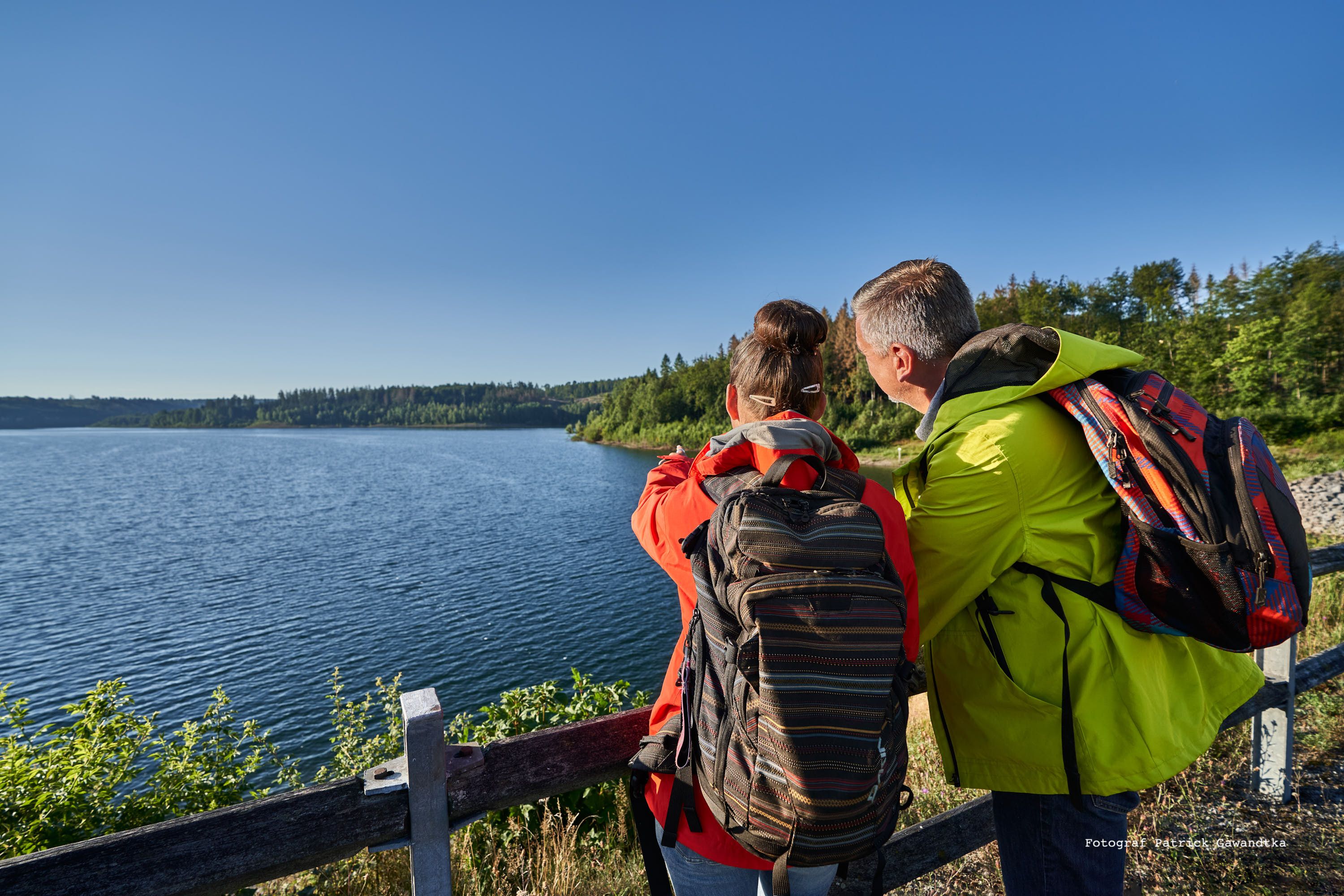 Zwei Personen mit Rucksäcken schauen auf einen See und bewaldete Uferlandschaft. Der Himmel ist klar und blau.