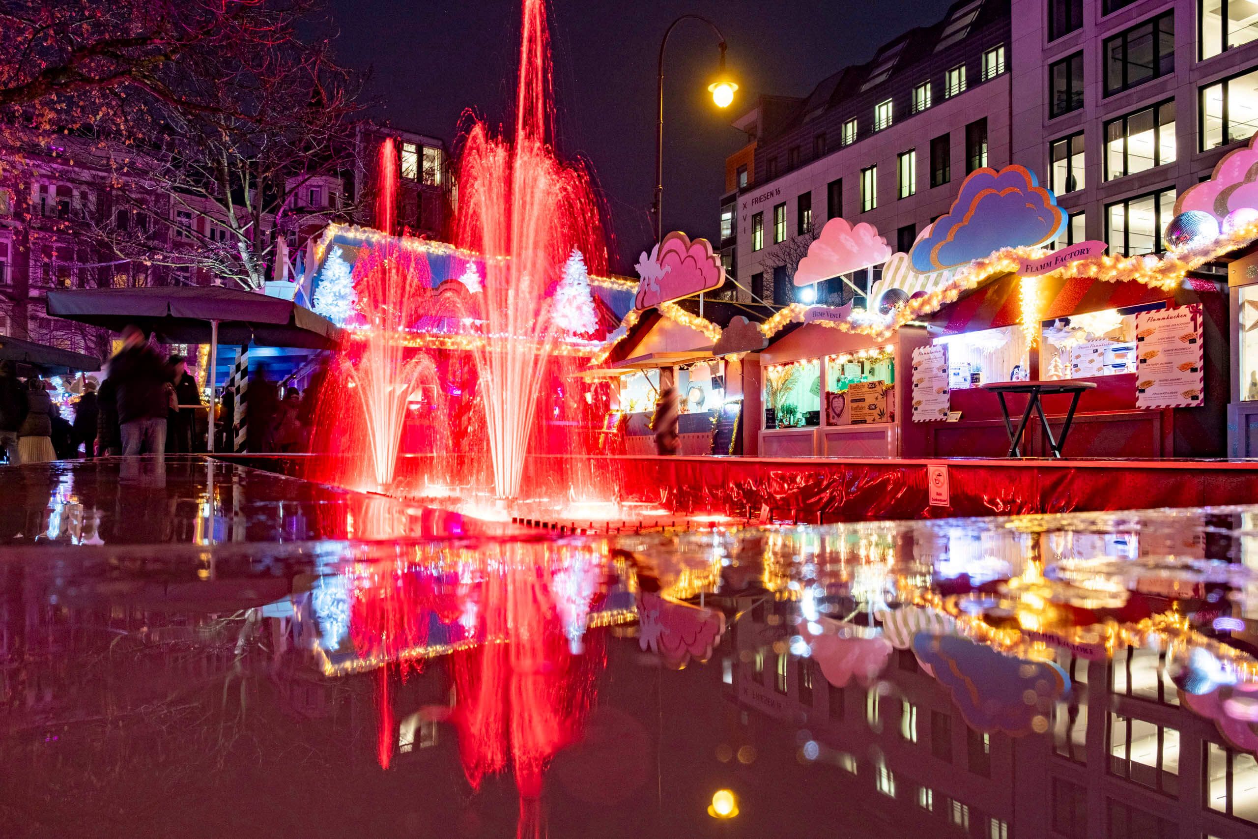 Bunte Weihnachtsmarktstände und ein beleuchteter Brunnen bei Nacht auf dem HEAVENUE Cologne.
