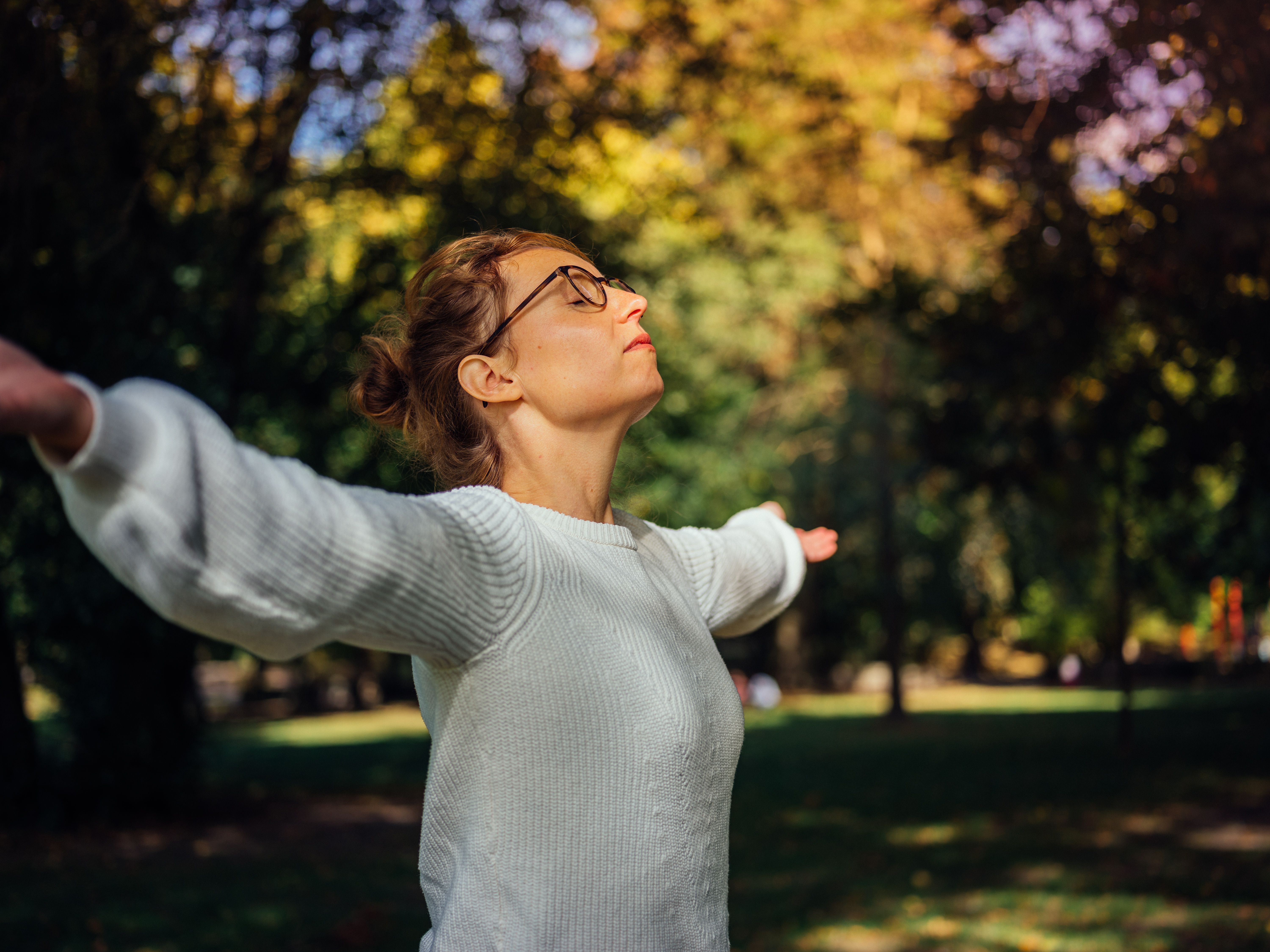 Frau in weißem Pullover steht mit ausgebreiteten Armen in einem sonnigen Park.