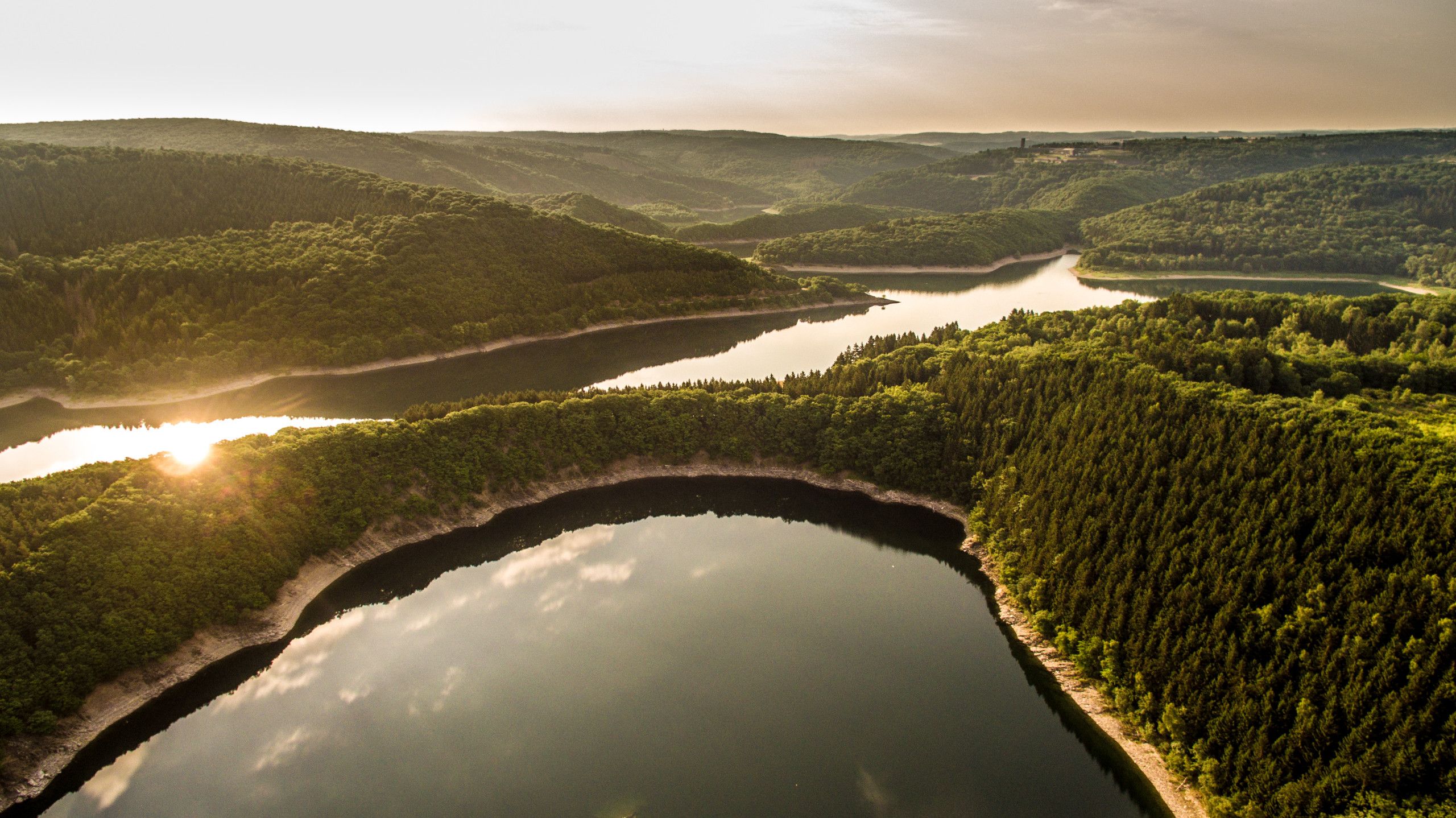 Luftaufnahme des Rursees mit bewaldeten Hügeln und Sonnenreflexion im Wasser. Die Sonne spiegelt sich im ruhigen Wasser wider.