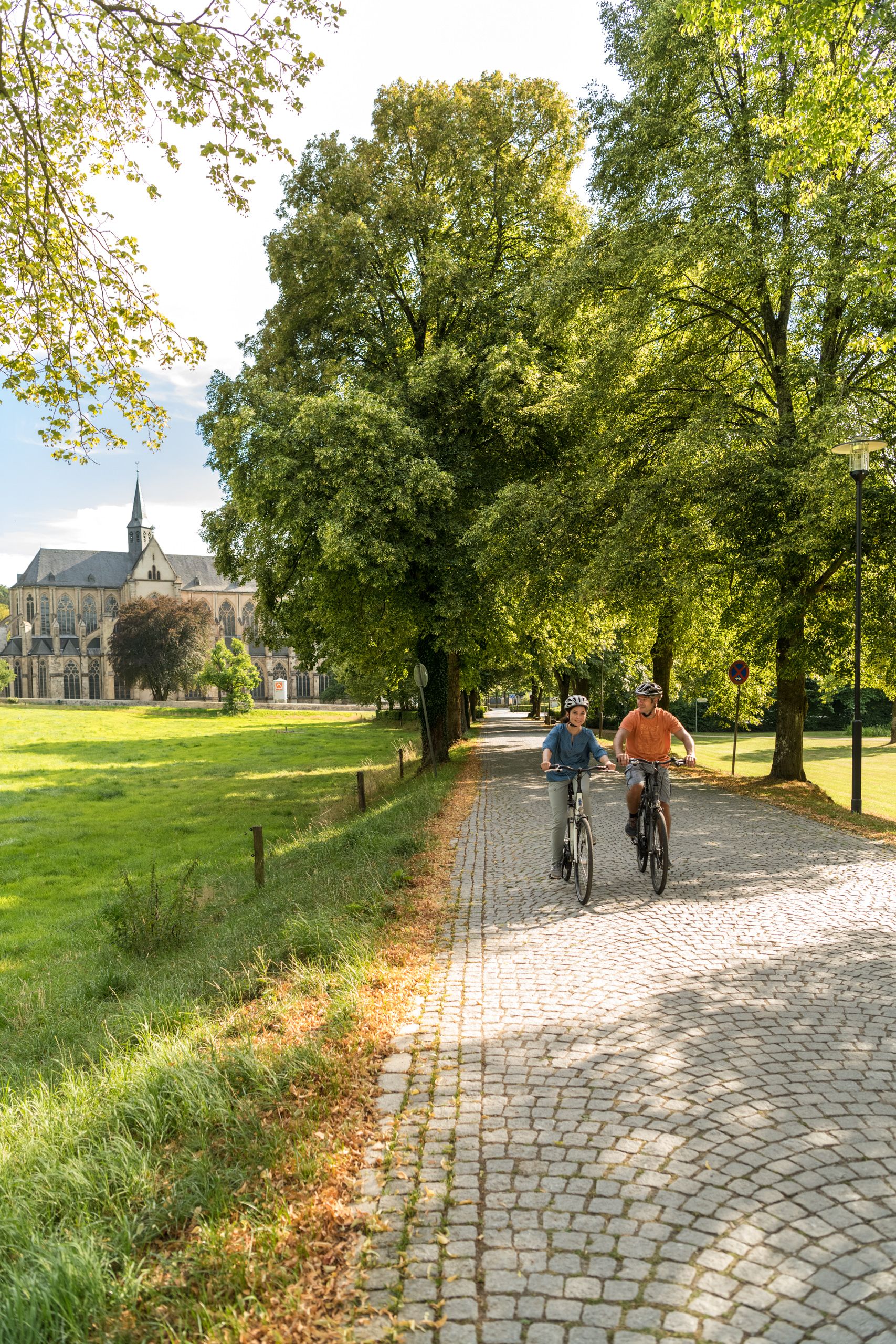Zwei Personen fahren auf einem gepflasterten Weg Fahrrad, im Hintergrund der Altenberger Dom und Bäume.