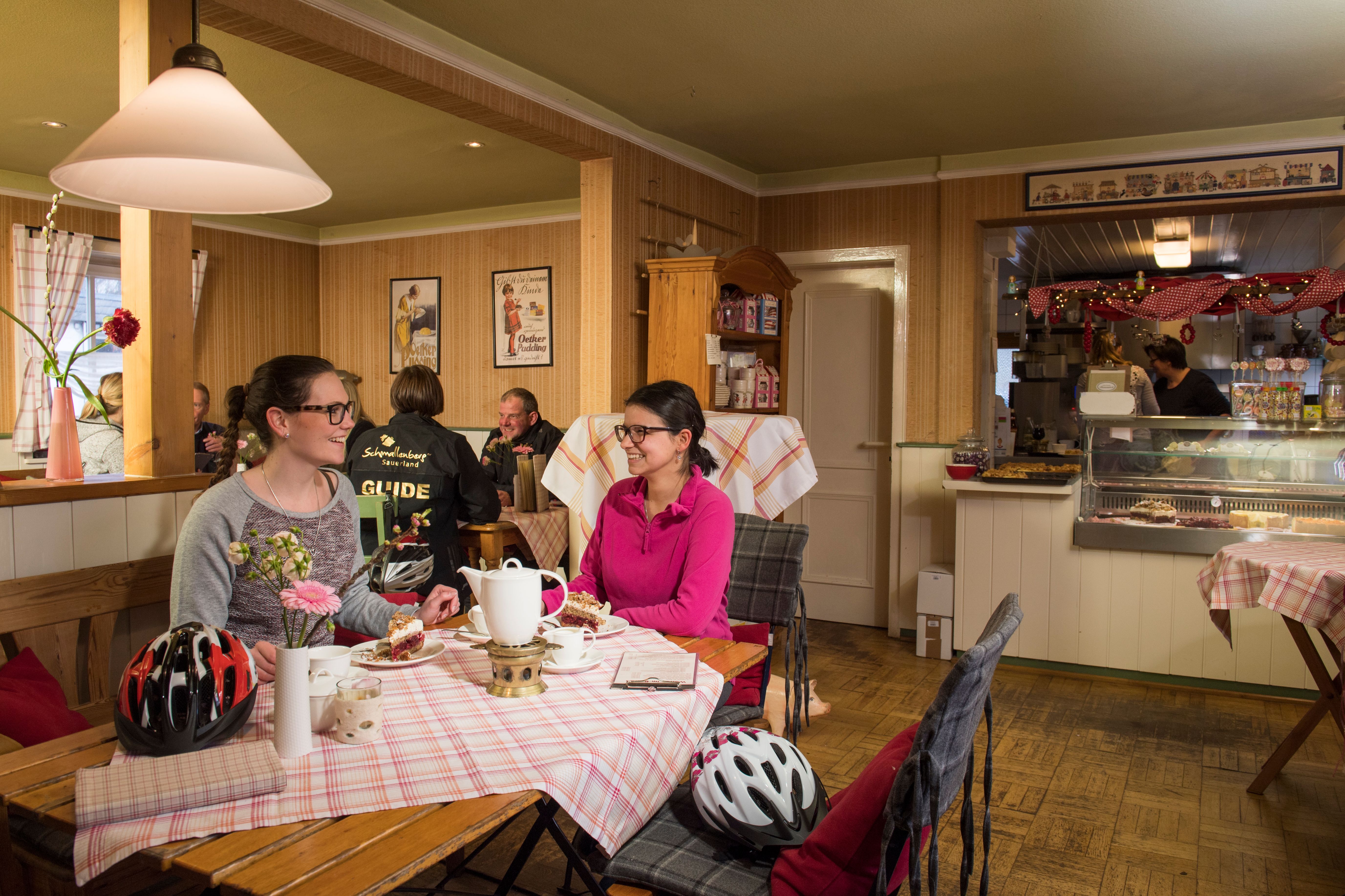 Zwei Frauen sitzen in einem Café, tragen Fahrradhelme und genießen Kuchen und Tee. Fahrradhelme liegen auf dem Tisch. Im Hintergrund sind weitere Gäste zu sehen.