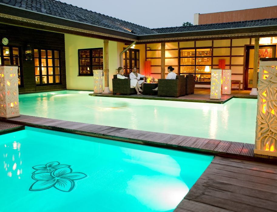 Zwei Personen in Bademänteln entspannen auf einer Terrasse neben einem beleuchteten Pool in der Bali Therme Bad Oeynhausen.