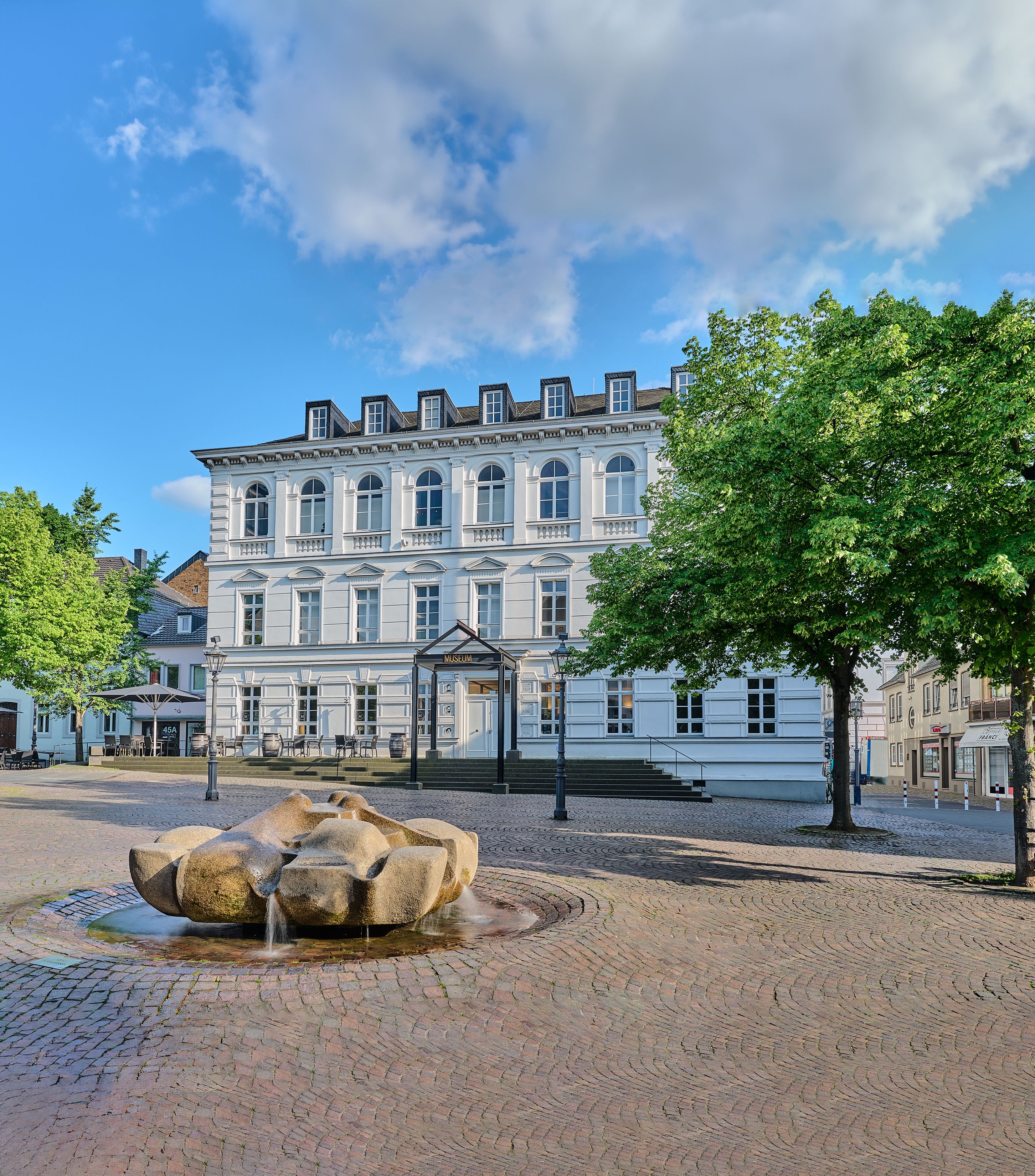 Siegburg Stadtmuseum mit Tourist Information: weißes historisches Gebäude am Platz mit Brunnen, Bäumen und blauem Himmel.