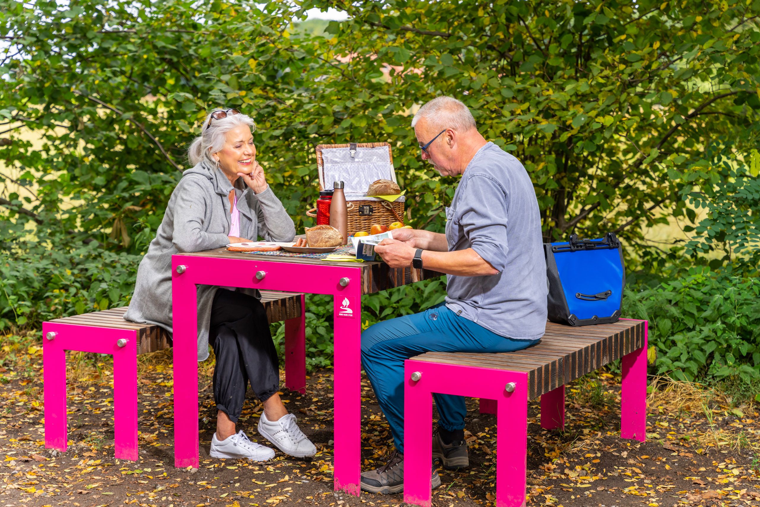 Ein älteres Paar sitzt an einem pinken Picknicktisch im Grünen und genießt eine Mahlzeit an der Bärenschleuse auf dem Hohe Mark Steig