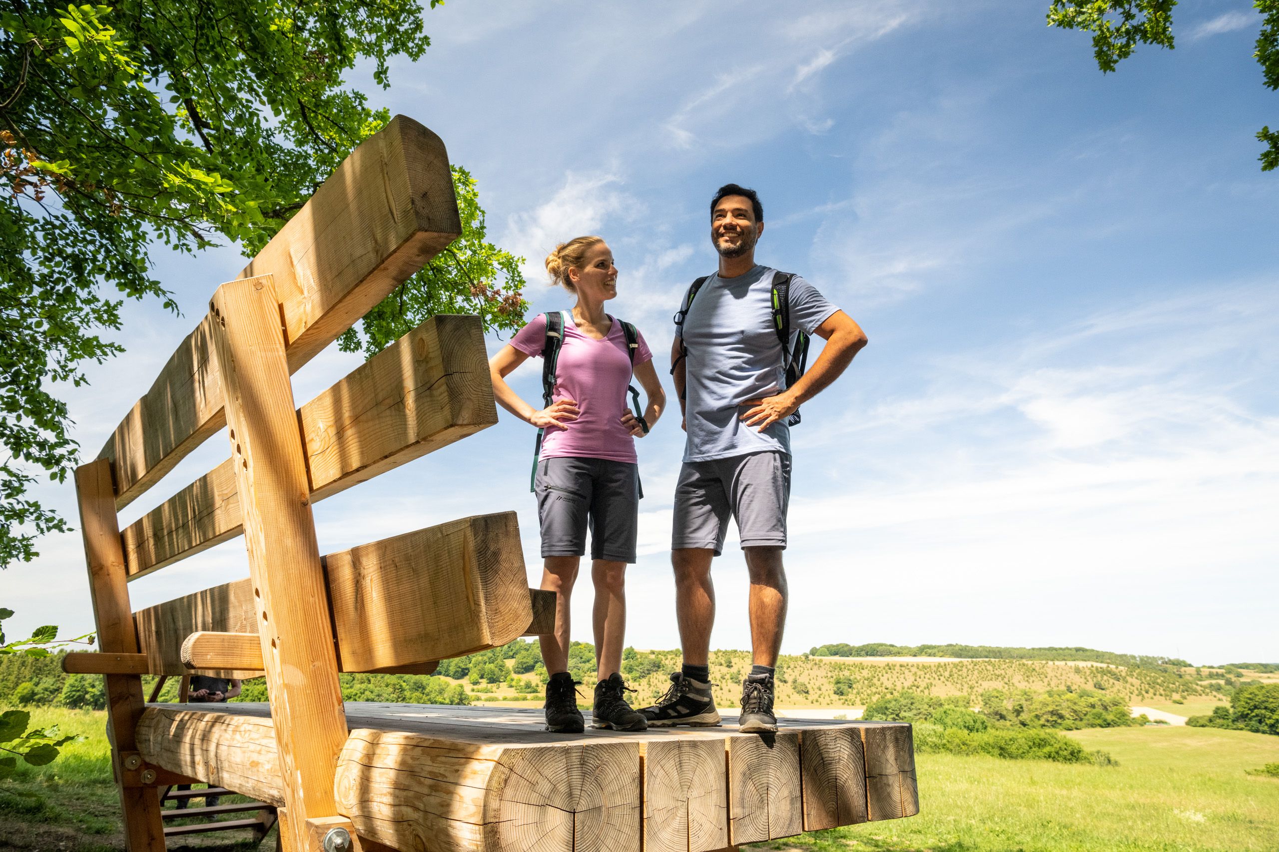 Zwei Personen stehen auf einer übergroßen Holzbank in einer grünen Landschaft unter blauem Himmel.
