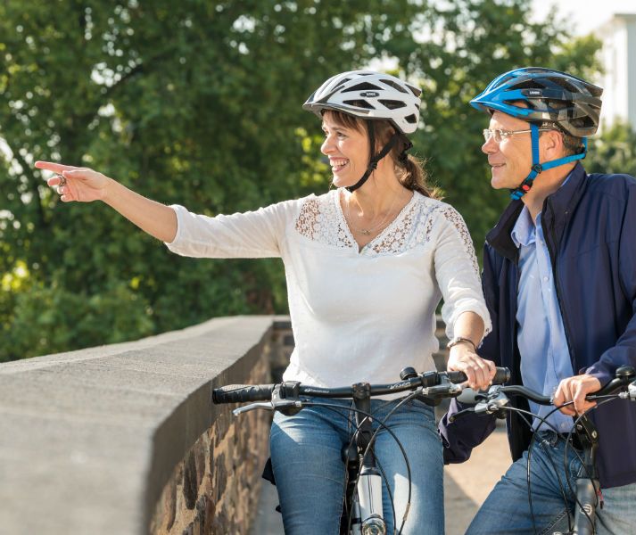 Zwei Personen mit Helmen auf Fahrrädern, eine zeigt lächelnd in die Ferne. Sie stehen auf einer Brücke mit grünem Hintergrund