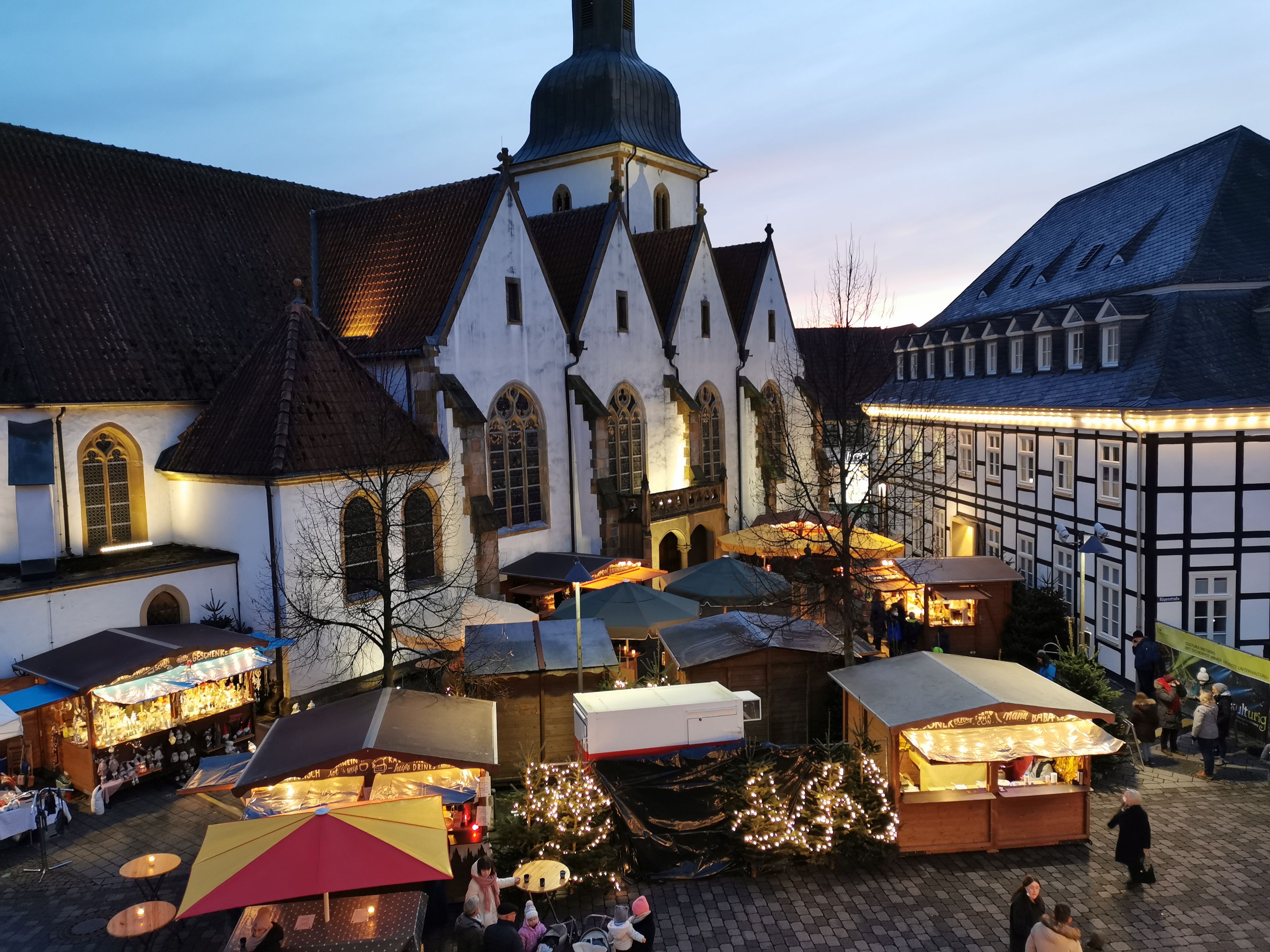Rietberger Adventsmarkt auf einem historischen Platz mit beleuchteten Holzhütten vor einer Kirche und Fachwerkhäusern in der Dämmerung.