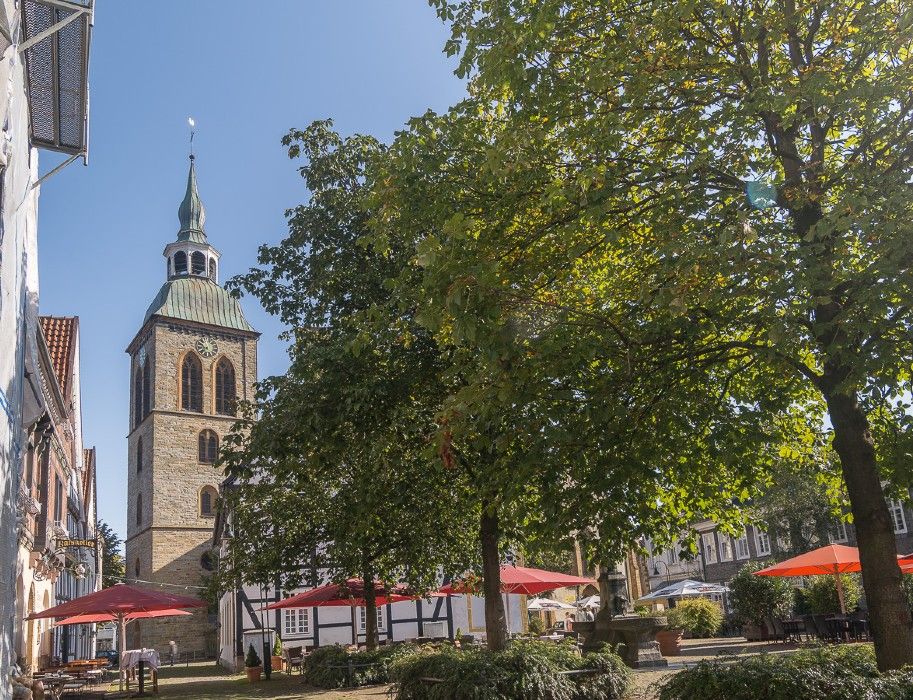Kirchturm in Rheda-Wiedenbrück mit Bäumen und Sonnenschirmen im Vordergrund.