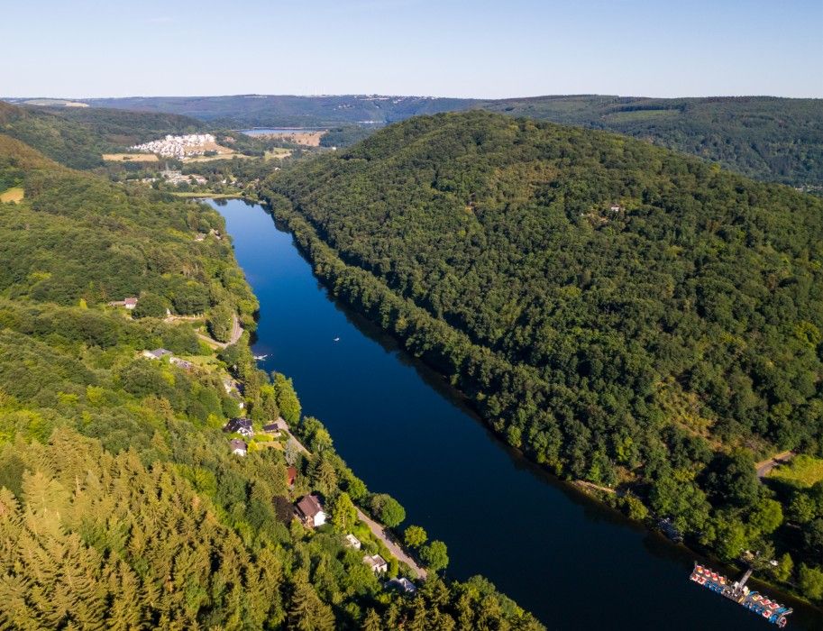 Luftaufnahme eines Flusses, der sich durch eine bewaldete Hügellandschaft schlängelt. Im Hintergrund sind Siedlungen und weite Wälder zu sehen.
