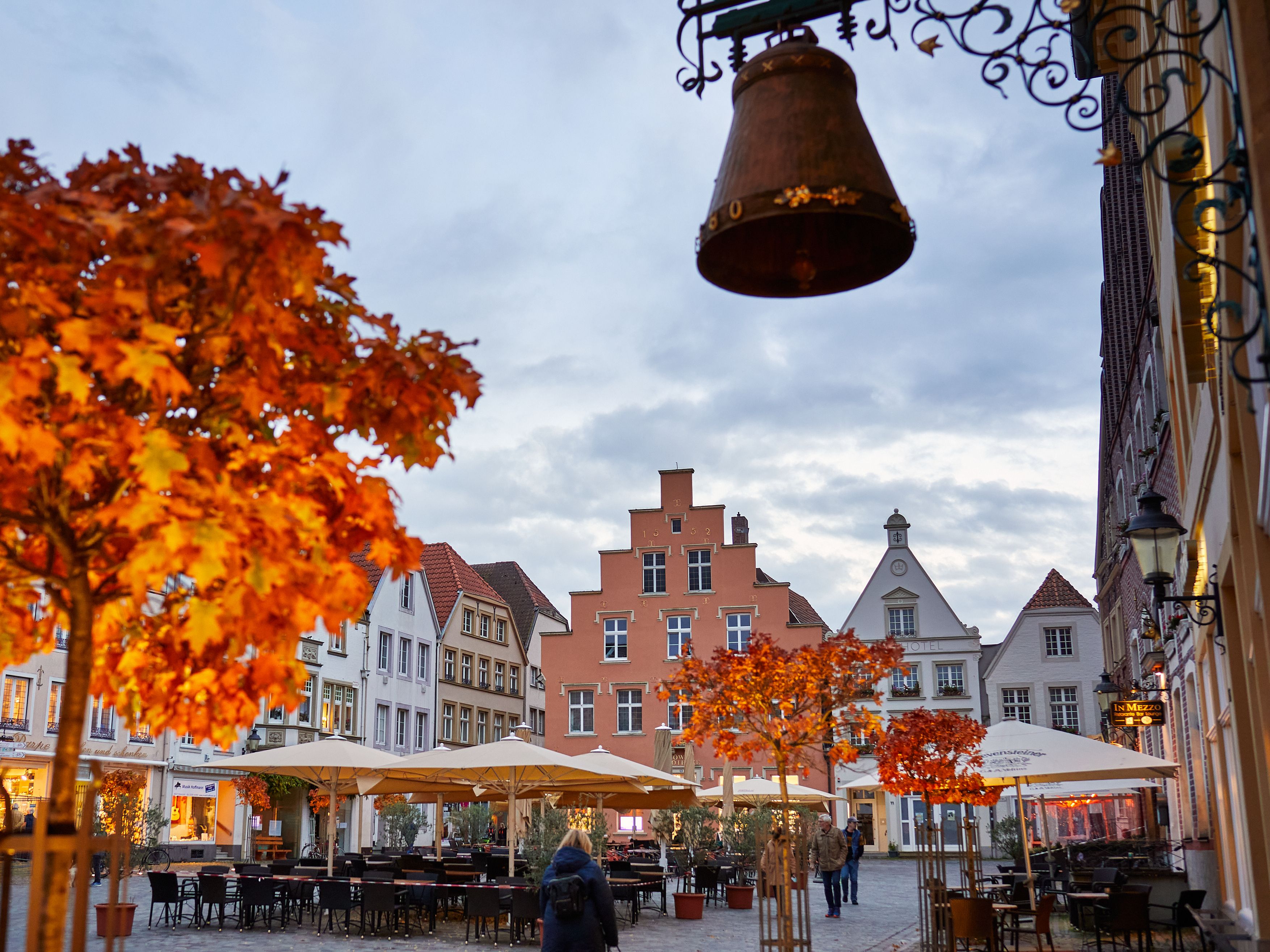 Herbstlicher Platz in Warendorf mit bunten Bäumen, historischen Gebäuden und einem Café mit Sonnenschirmen. Eine Glocke hängt im Vordergrund.