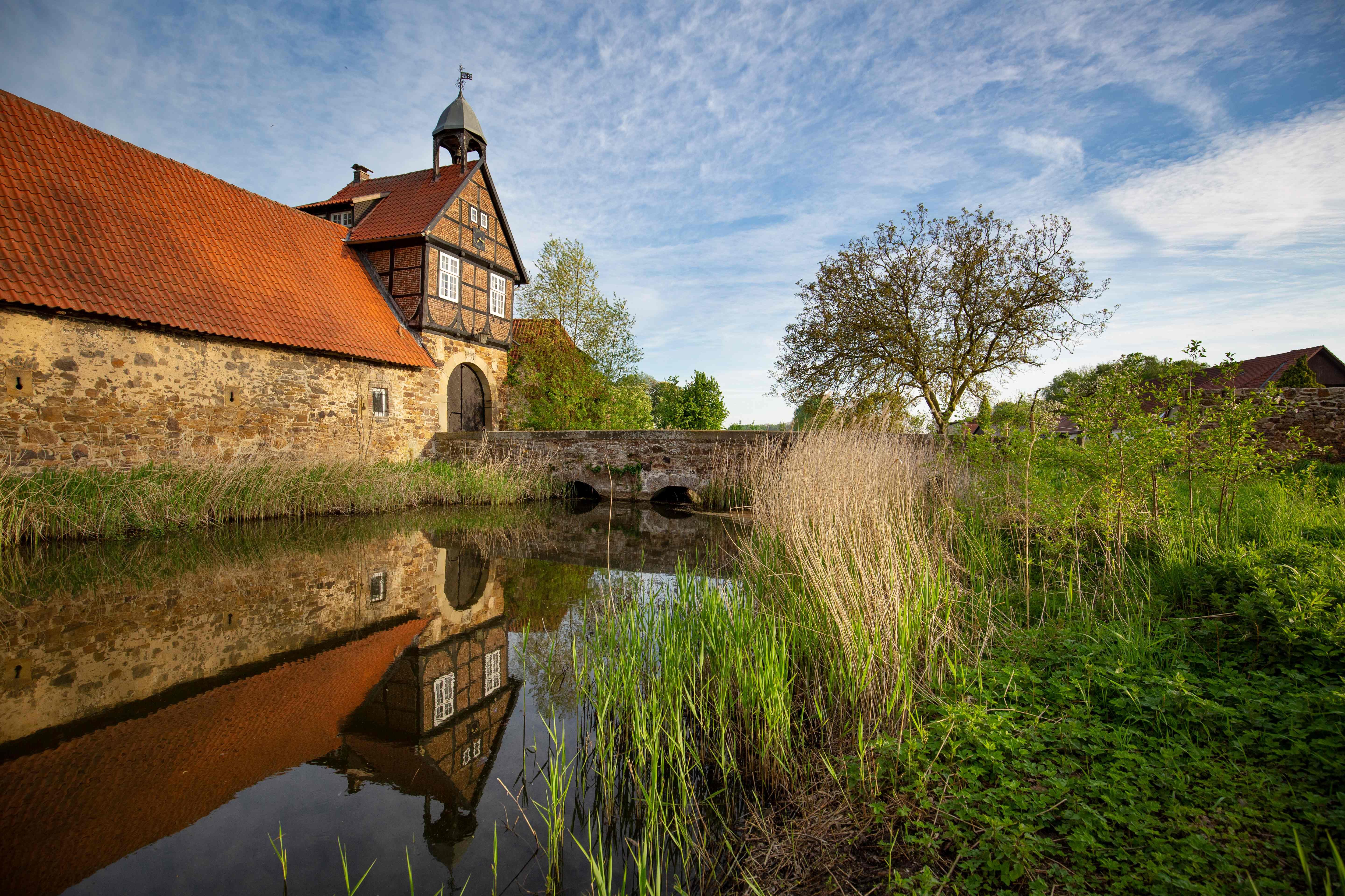 Historisches Gebäude mit Fachwerk und rotem Dach, reflektiert in einem Wassergraben, umgeben von grüner Vegetation und blauem Himmel.