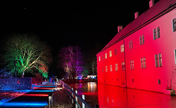 Schloss Benkhausen bei Nacht, beleuchtet in bunten Farben mit einem beleuchteten Weg und Bäumen im Hintergrund.